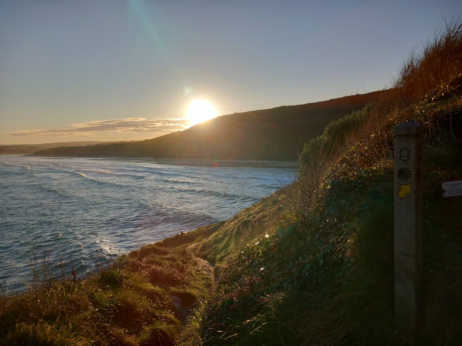 A scenic coastal view at sunset shows the sun partially obscured by headlands on the left. The ocean waves roll toward the shore on the right. A trail signpost with directional arrows stands on a grassy hillside, leading the eye toward the shimmering water.