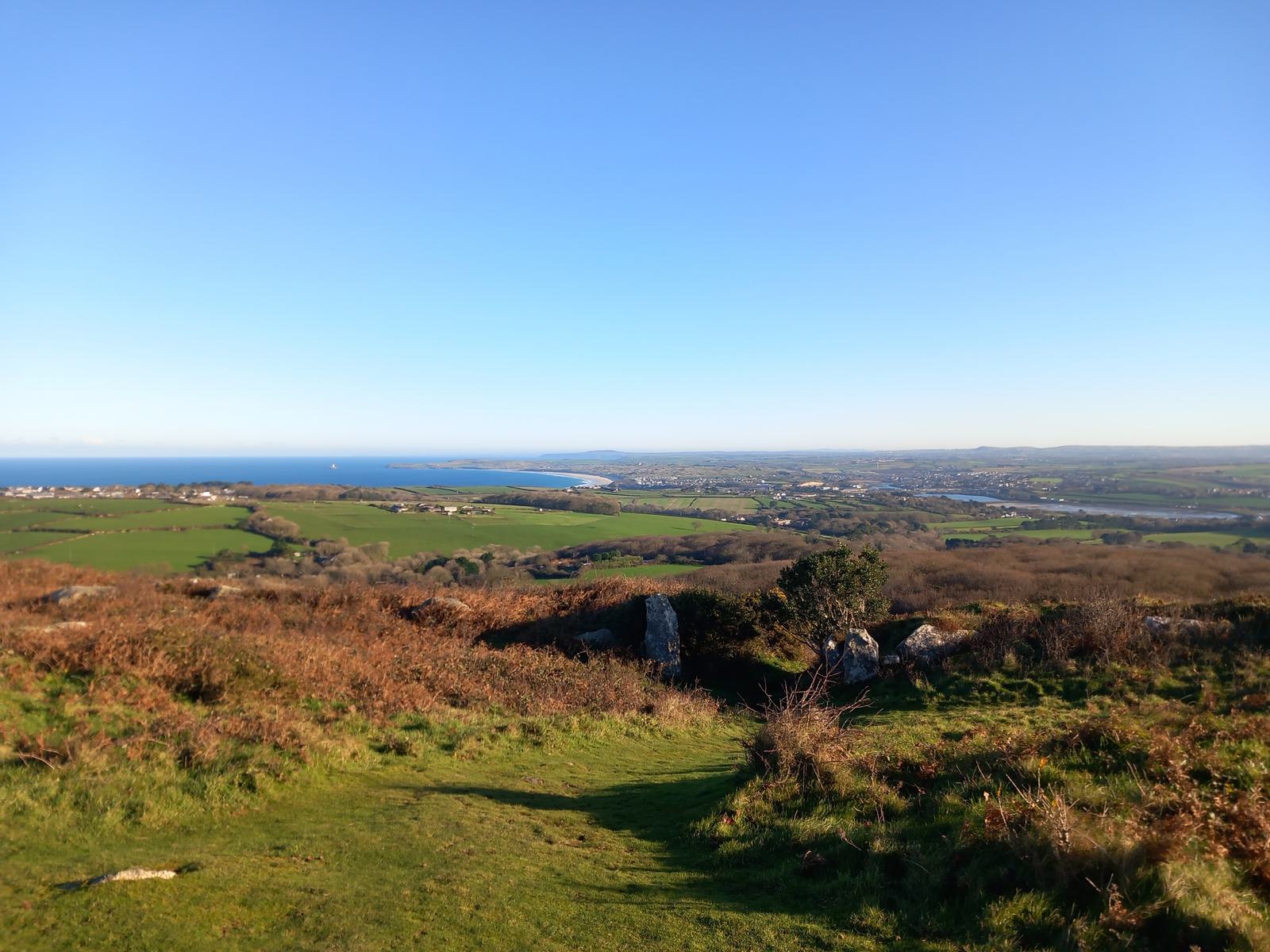 A scenic landscape featuring a vast green field with patches of brown foliage, leading to a distant coastline under a clear blue sky. The horizon separates the bright sea from the expansive land dotted with small structures and rolling hills.