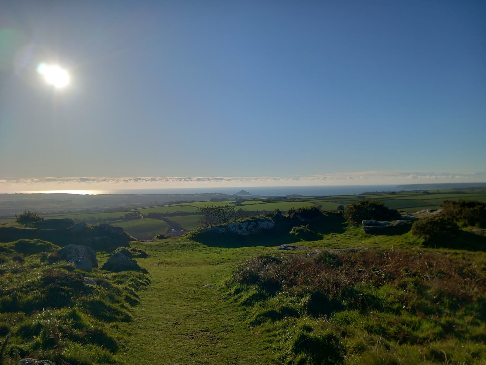 A scenic landscape view of a grassy hilltop under a clear blue sky with the sun shining brightly. In the distance, there are green fields, a body of water, and a faint outline of a mountainous area on the horizon. Rocks and bushes are scattered in the foreground.