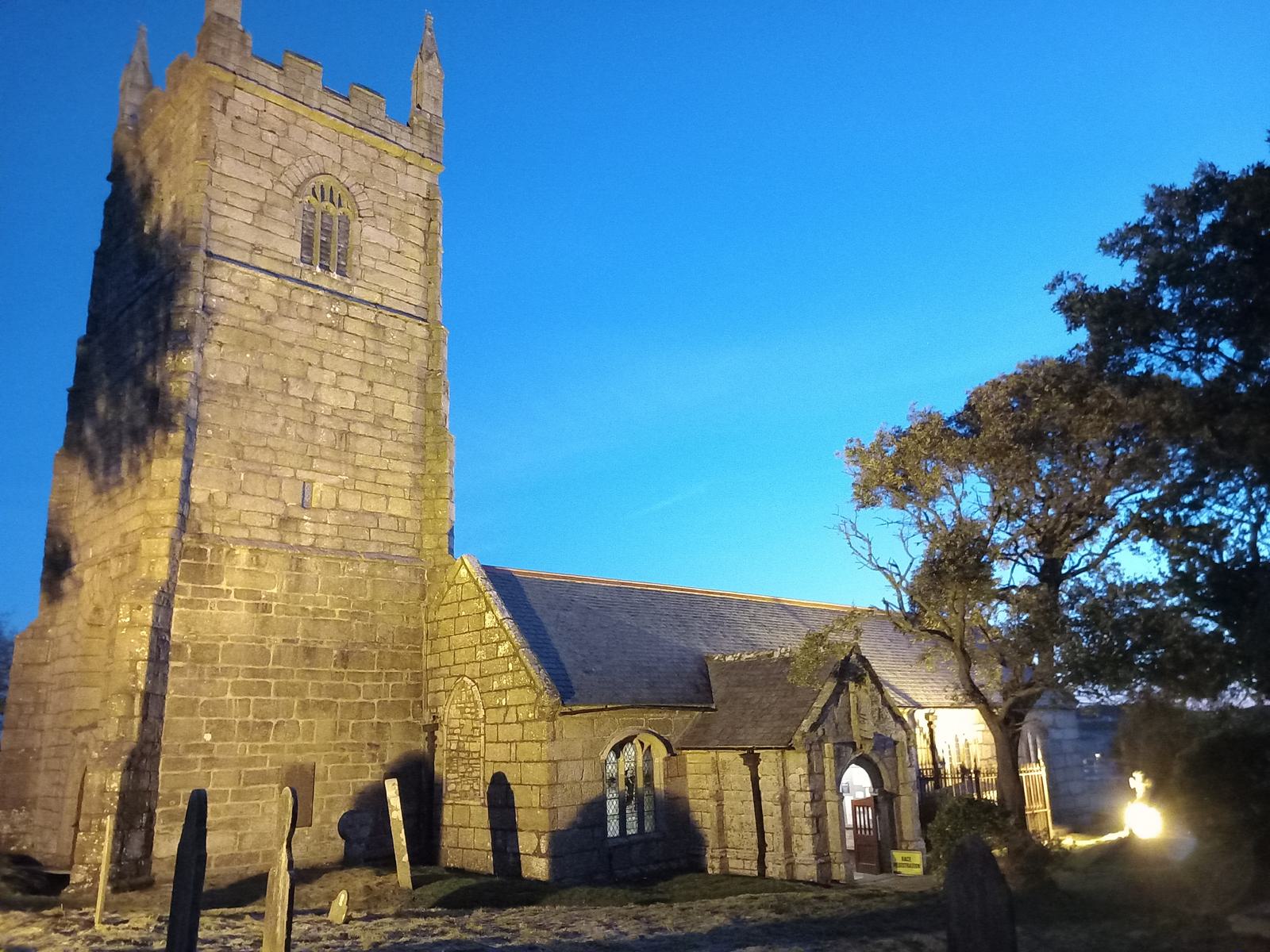 A stone church with a tall, square clock tower stands illuminated against a twilight sky. The church has a small annex, brightly lit, and is surrounded by several old gravestones. Two trees frame the right side of the scene.