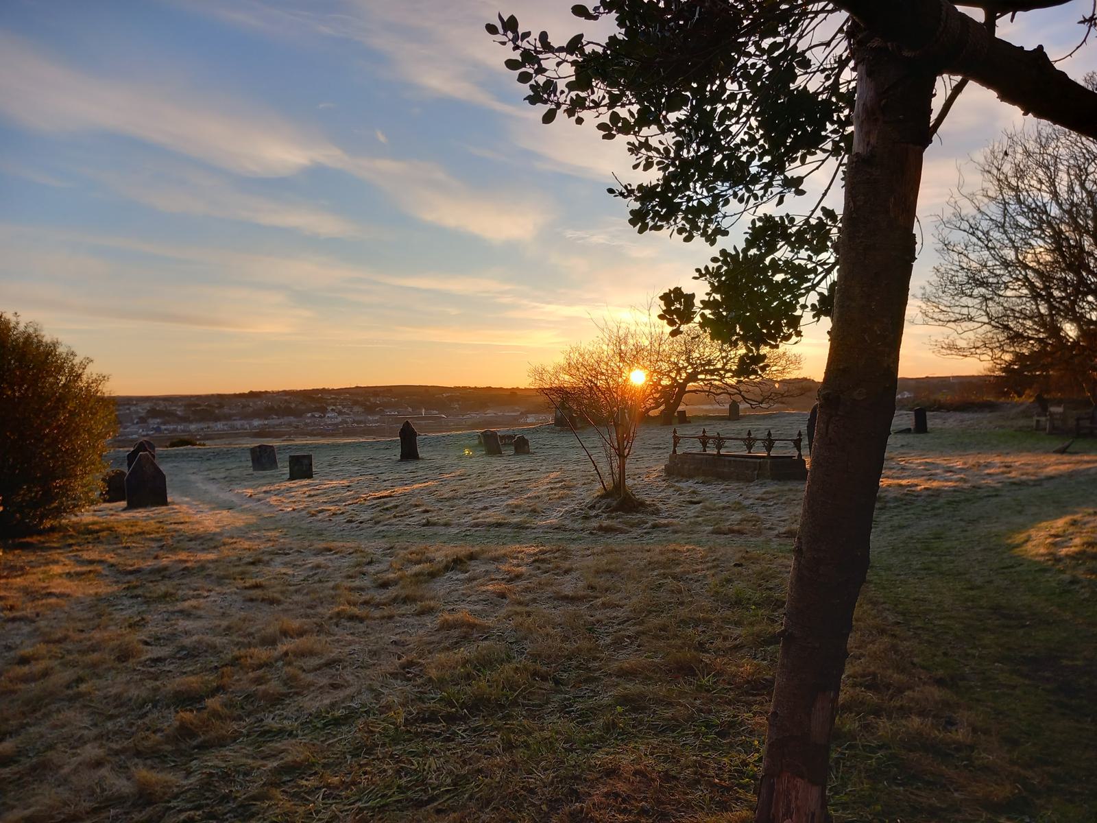 A serene sunset over a frosty graveyard, with sunlight peeking through sparse tree branches. The sky is painted with hues of orange and blue. Tombstones and statues are scattered across the ground, casting long shadows on the frost-covered grass.