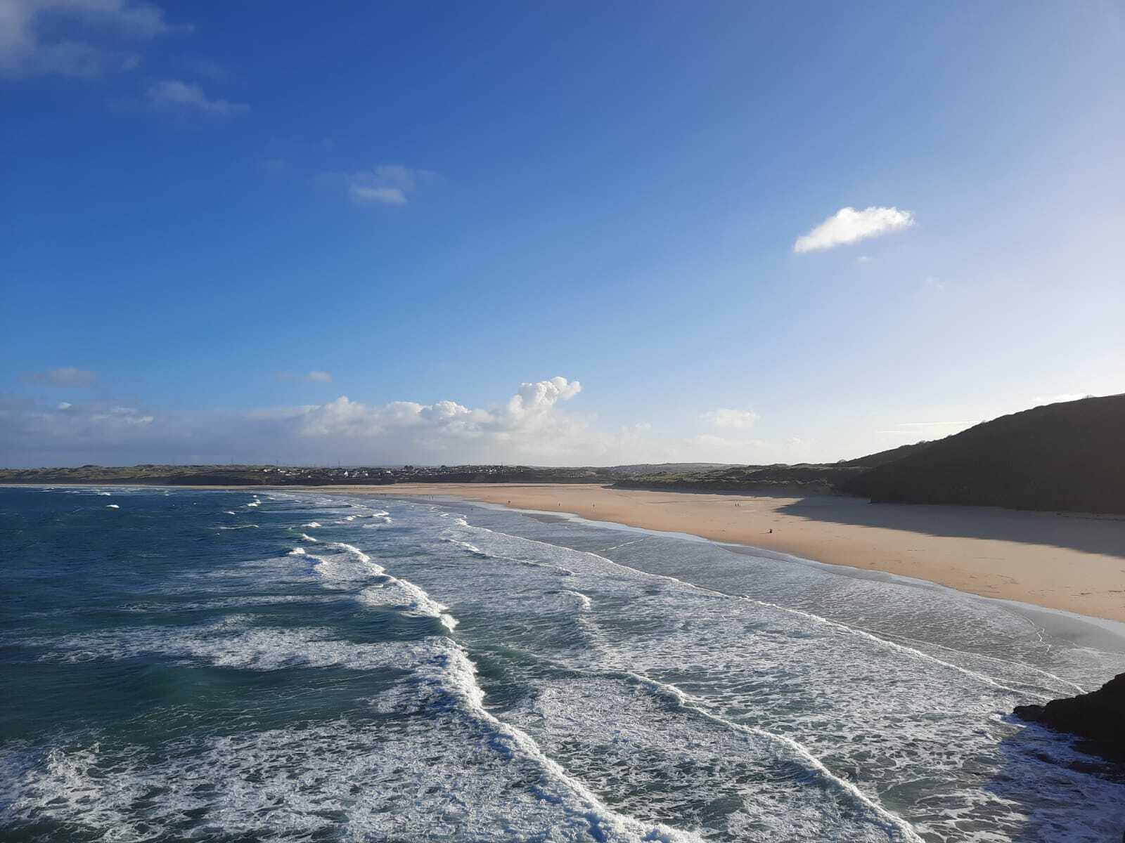A scenic view of a sandy beach against clear blue skies and gentle waves rolling onto the shore. The beach curves along the coastline, bordered by hills and distant buildings. The overall setting is tranquil and sunlit.