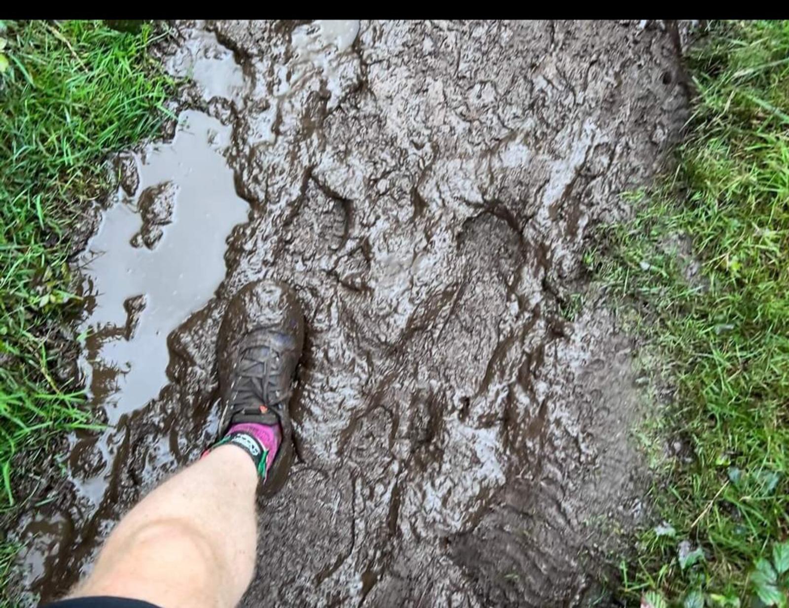 A close-up view of a person's muddy foot and leg, wearing a running shoe and a sock with colorful designs, stepping into a waterlogged, muddy path surrounded by patches of green grass. The scene suggests a challenging outdoor activity in wet conditions.