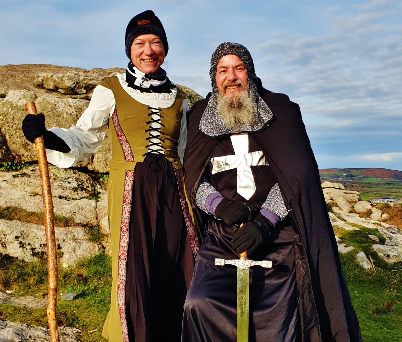 Two people in medieval costumes stand outdoors on a rocky landscape. The person on the left is in a brown dress with a white blouse and black hat, holding a wooden staff. The person on the right is in a black knight's outfit with a cross emblem, holding a sword.