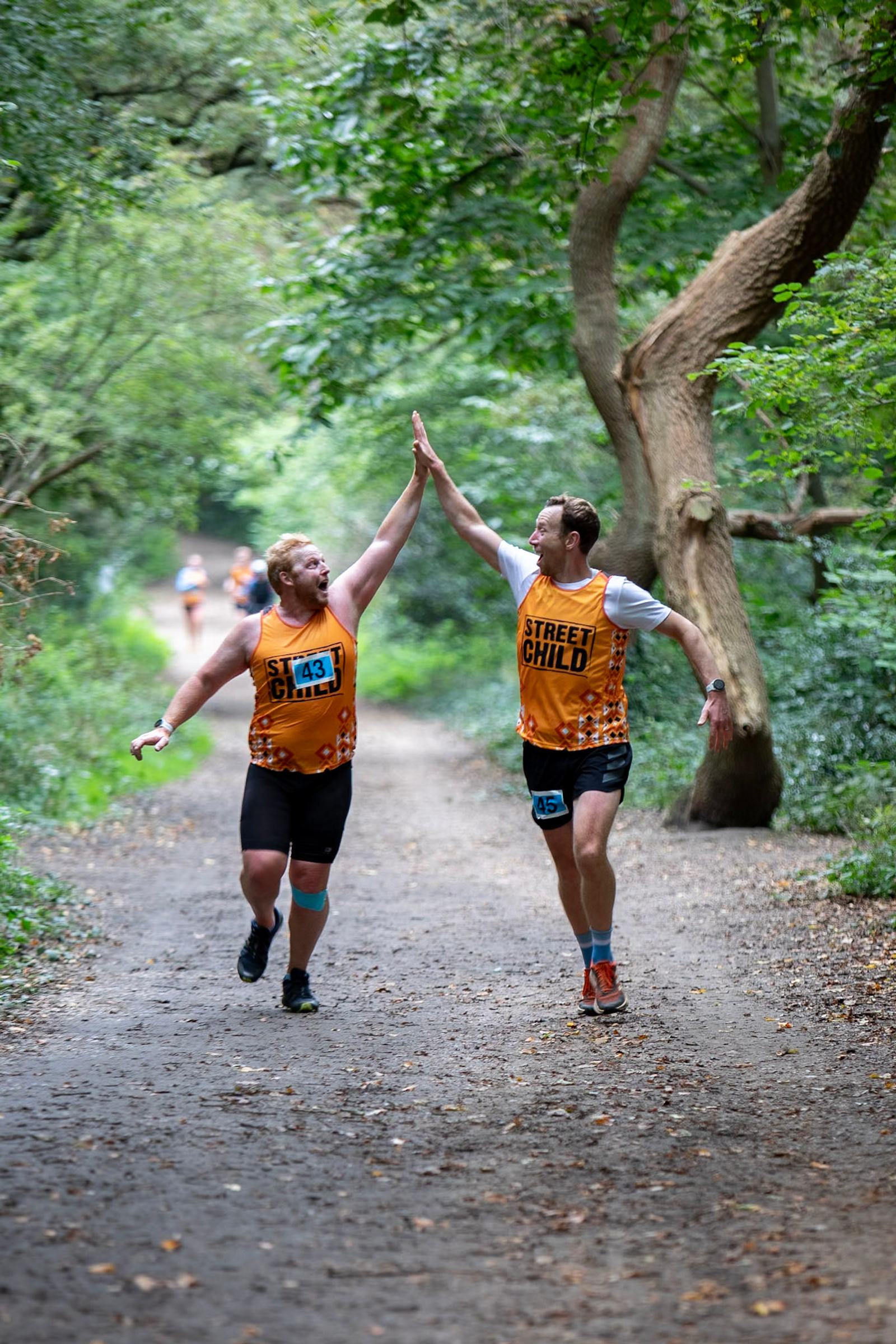 Two runners in orange jerseys high-five each other on a forest trail. They appear happy and energetic, with race bibs on their shirts. The surrounding area is lush with green trees and foliage.