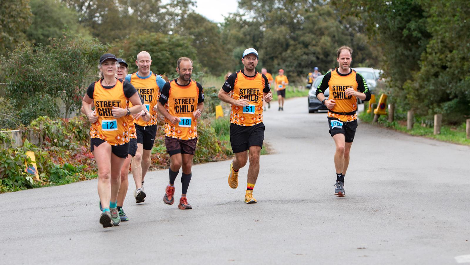 Five runners wearing matching orange "Street Child" shirts participate in a race on a wooded road. They appear focused and engaged, with some smiling. The background features trees and a parked car.