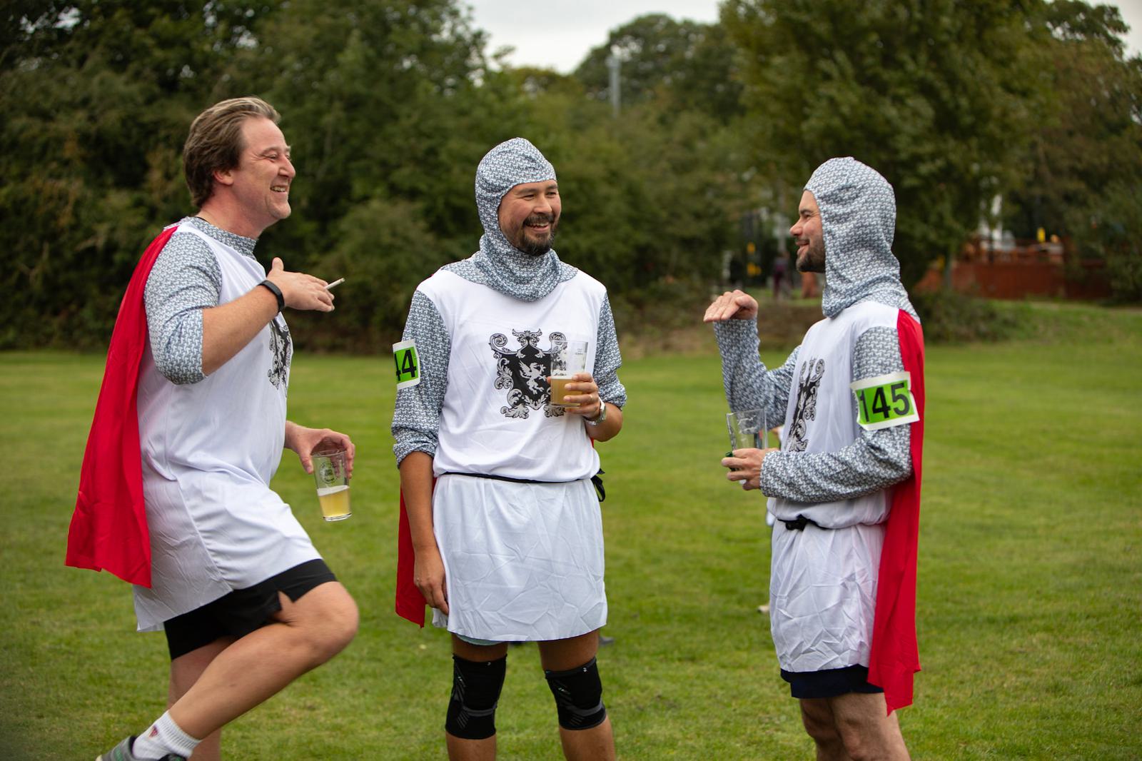 Three people in medieval knight costumes laugh and chat outdoors on a grassy field. They wear chainmail hoods, white tunics with black heraldic symbols, red capes, and numbered armbands. Two hold plastic cups. Trees are visible in the background.