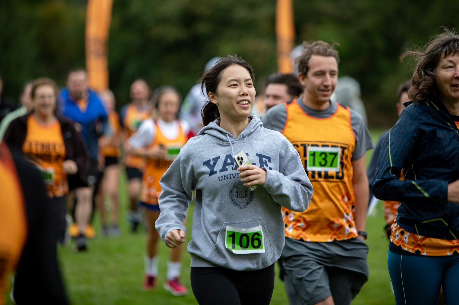 A woman wearing a Yale hoodie and a green race bib labeled "106" jogs in a group of people. Some are wearing orange and black team vests with numbers "137" and "149." The runners are outdoors on grass, surrounded by blurred greenery.