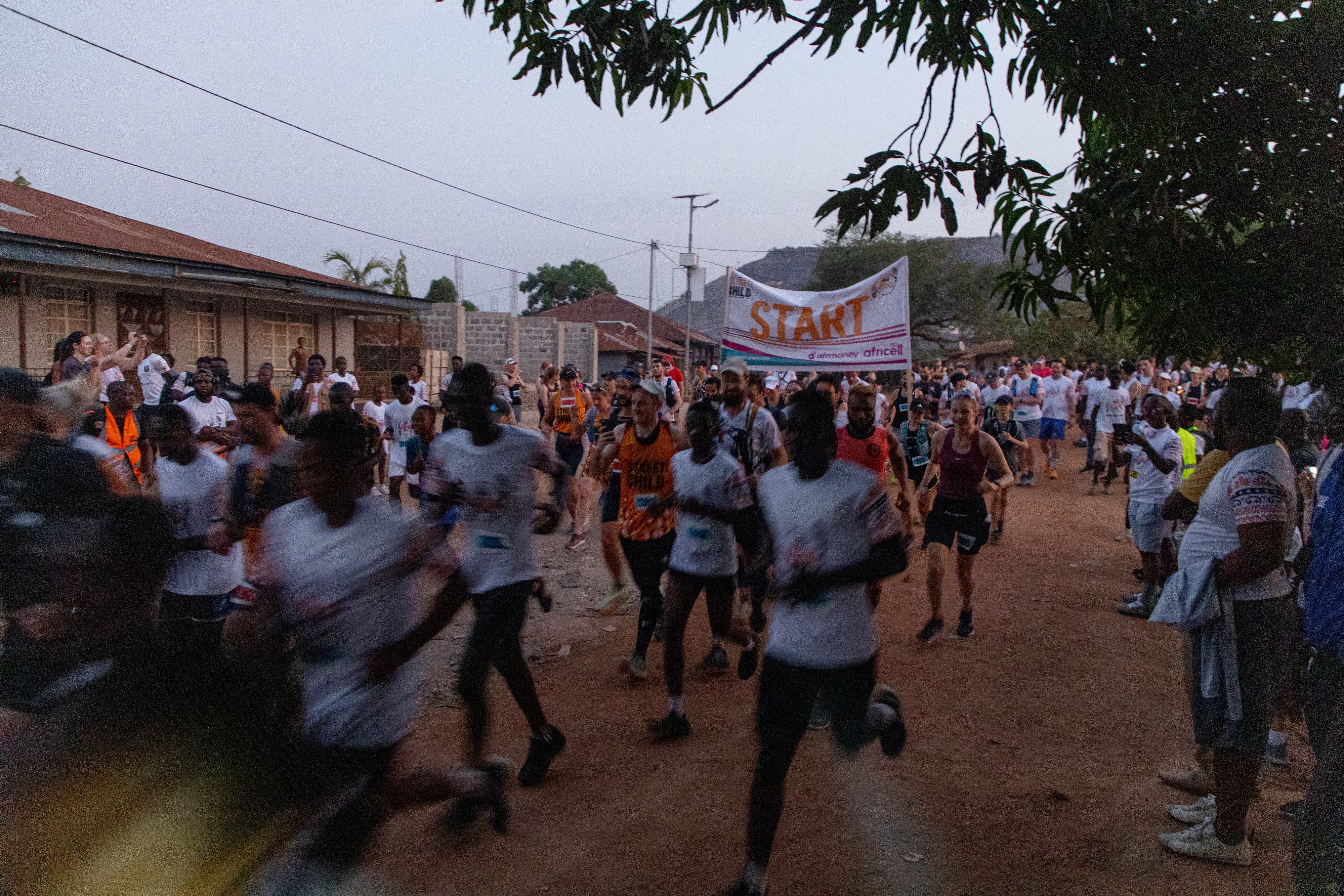 A large group of runners participate in a race on a dirt road, passing under a "START" banner. Onlookers and other participants are visible in the background, with buildings and trees lining the route. The sky appears overcast.