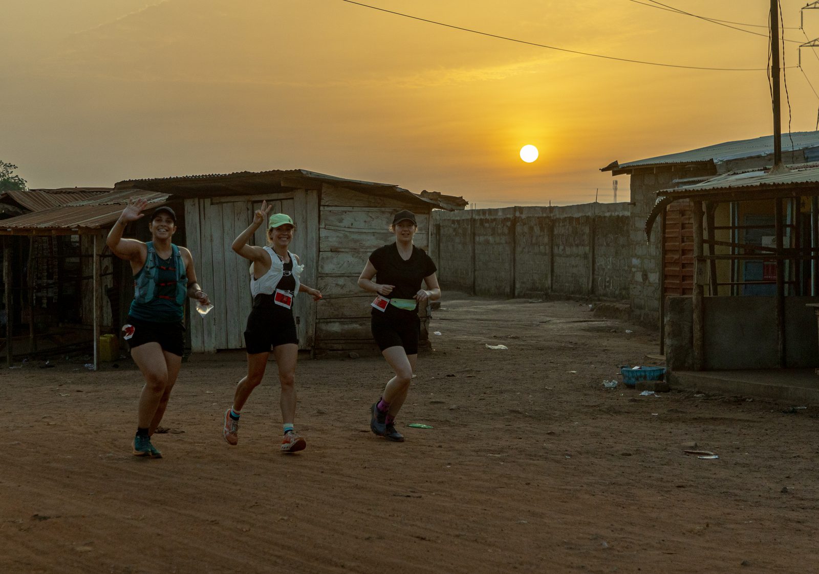 Three individuals are running on a dirt road at sunrise, passing through an area with small wooden and metal-roofed structures. They appear joyful, with one person waving at the camera. They are dressed in athletic gear and wear bibs, suggesting participation in a race.