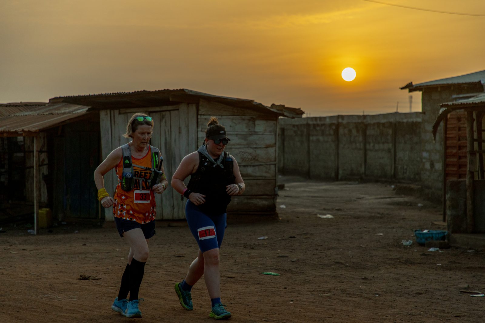 Two individuals, wearing athletic gear and bibs, are running on a dusty road near rustic wooden buildings during sunrise or sunset. The sky is orange, and the sun is low on the horizon. The setting appears to be a rural or semi-urban area.