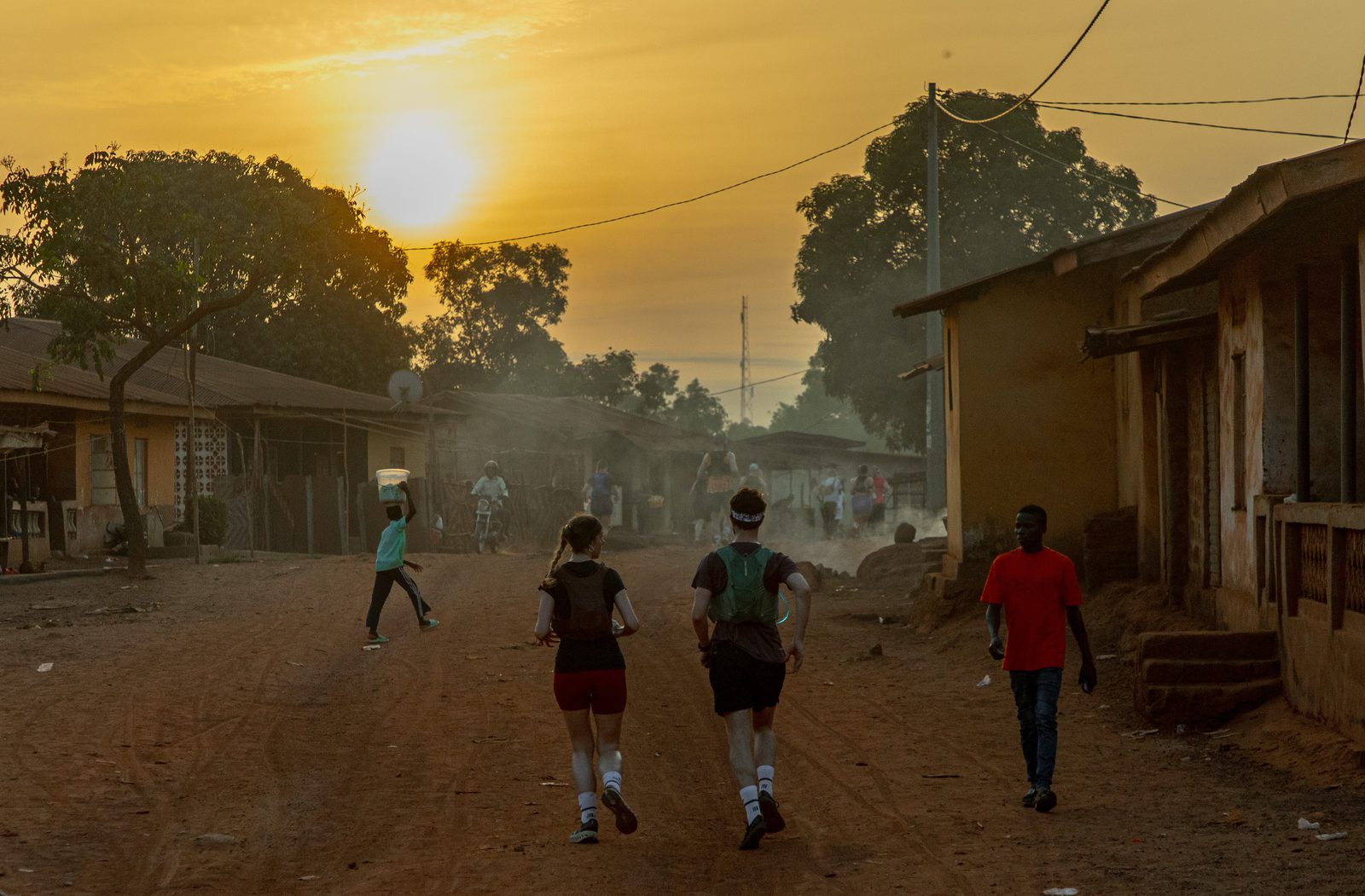 Two people jog down a dirt road through a village at sunrise, passing locals walking along the street. Low, single-story buildings and trees line the road, with a hazy, golden sky in the background.