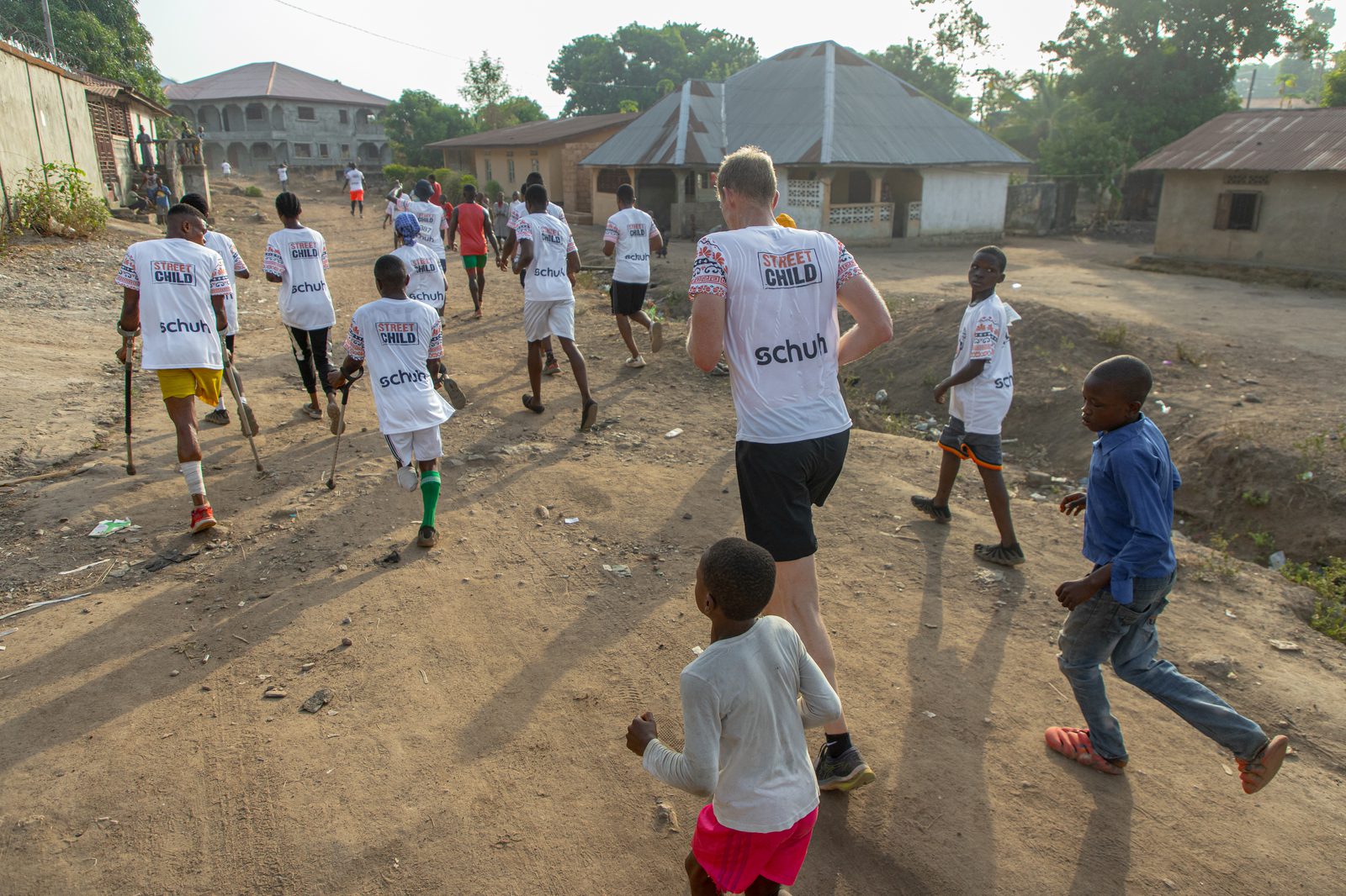 A group of people, including children and adults wearing matching "Street Child" shirts, are running along a dirt road in a rural area with sunlit trees and buildings in the background. Some local children join them, wearing casual clothing and no shirts.