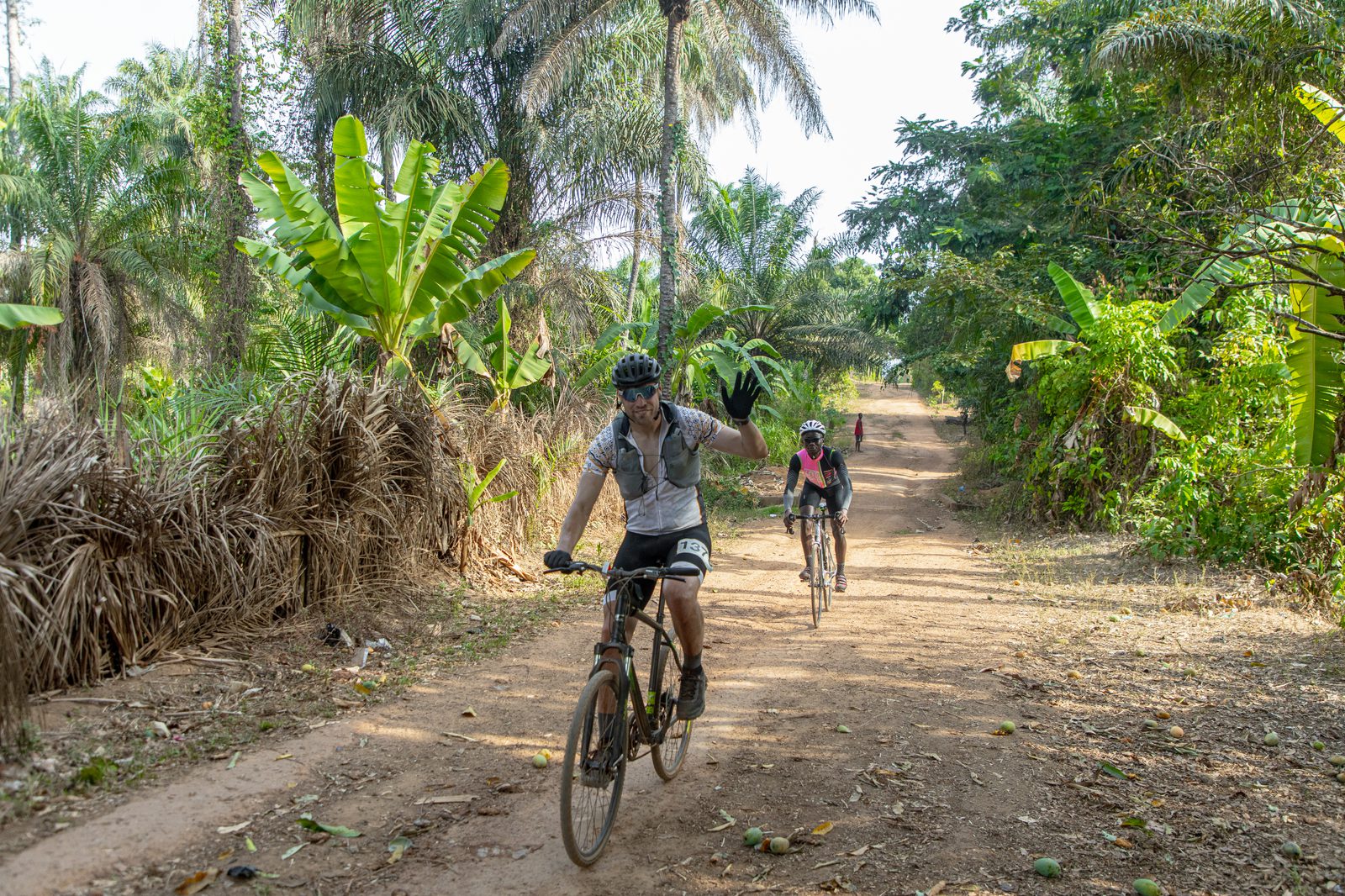Two cyclists ride on a dirt path through a lush, tropical landscape with tall palm trees and green vegetation. The lead cyclist is waving and wearing a helmet, sunglasses, and a grey shirt. The trailing cyclist is slightly out of focus. It's a sunny day.