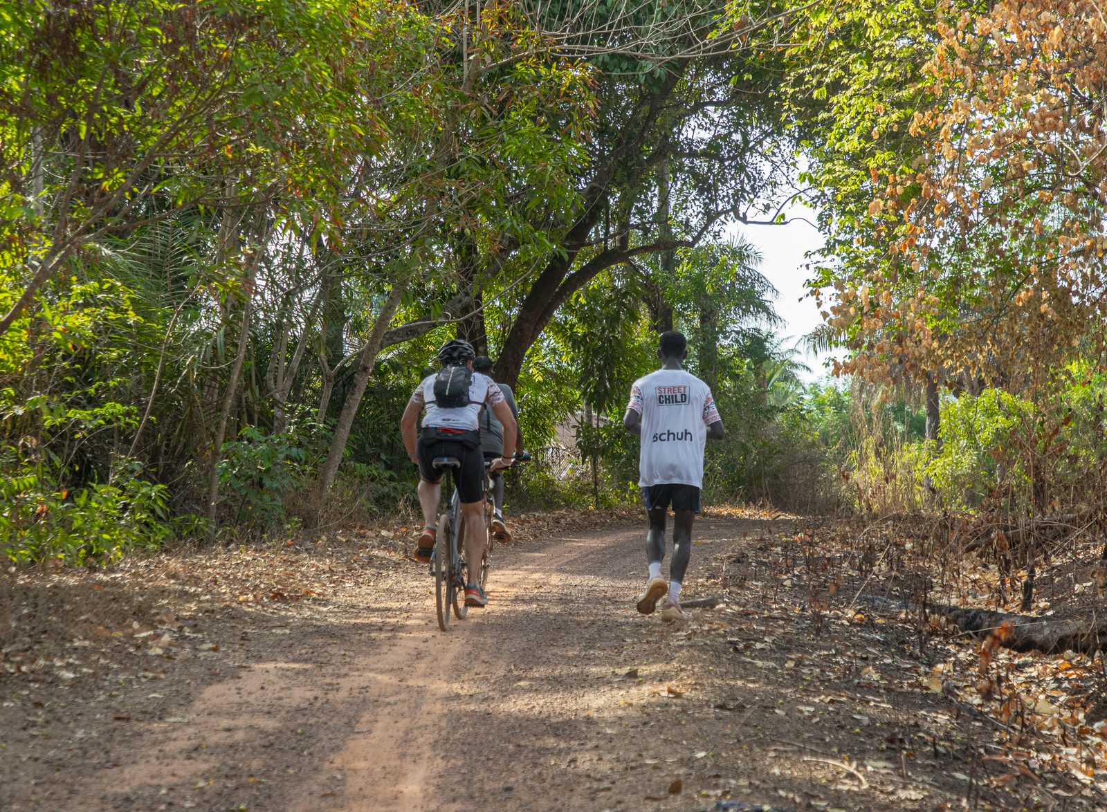 Two people are seen from behind on a dirt path surrounded by lush greenery. One is cycling, and the other is running. Both wear white shirts printed with text, and the path is partly shaded by overhanging branches. Dappled sunlight filters through the leaves.