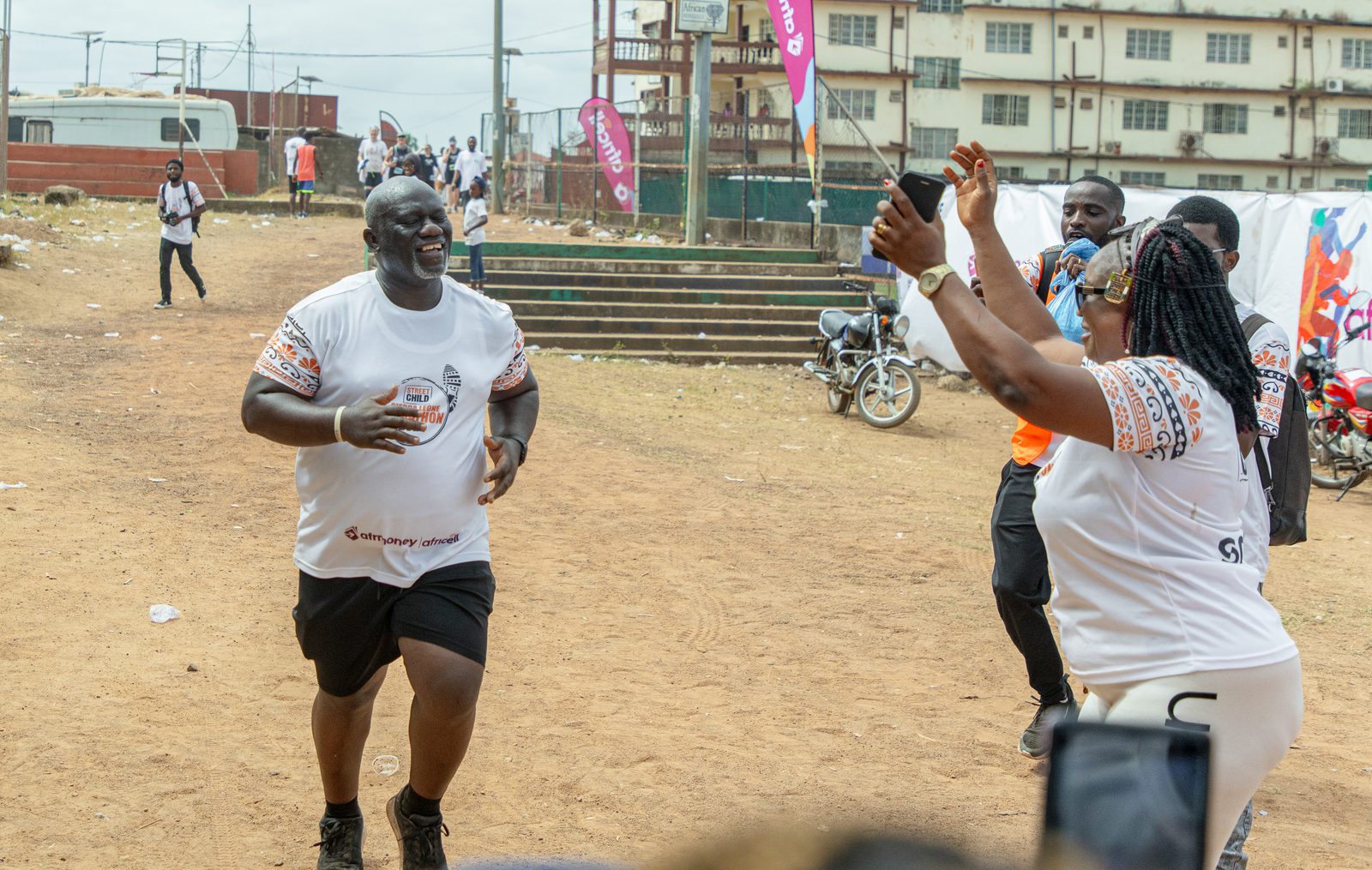 A man in a white athletic shirt and black shorts runs towards a cheering woman, who is taking his picture with her phone. They are outdoors on a dirt field, with several other people and buildings in the background. The atmosphere is festive and supportive.
