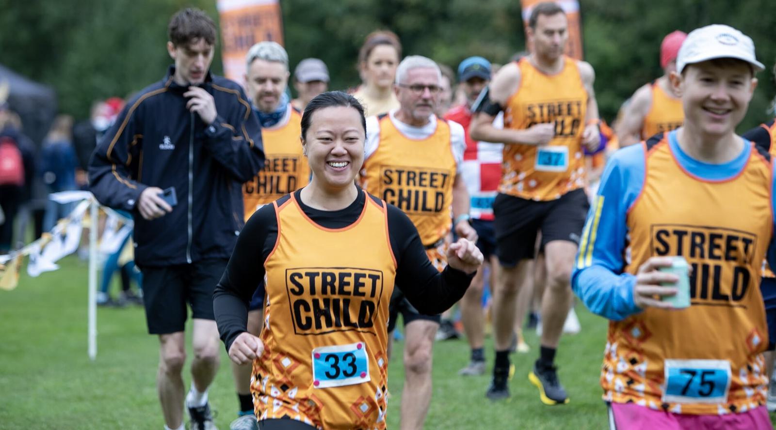 Runners participating in a race, wearing orange "Street Child" vests and numbered bibs, race along a grassy path. The participants are of various ages, with trees in the background and a festive atmosphere.