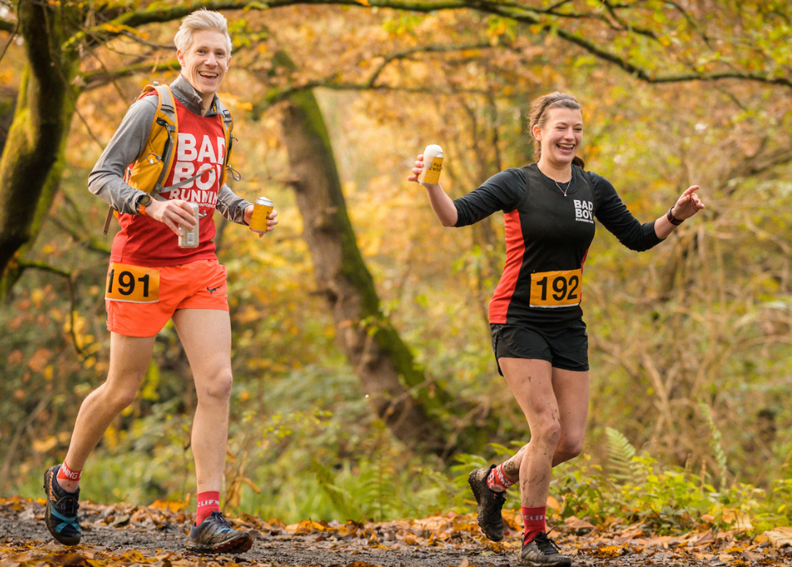 Two runners, wearing numbered bibs 191 and 192, smile and hold drinks in a wooded outdoor setting. The person on the left is in a red outfit, while the person on the right wears black and red. Logos for "Street Child Craft Half" and "LOCAL" appear below them.