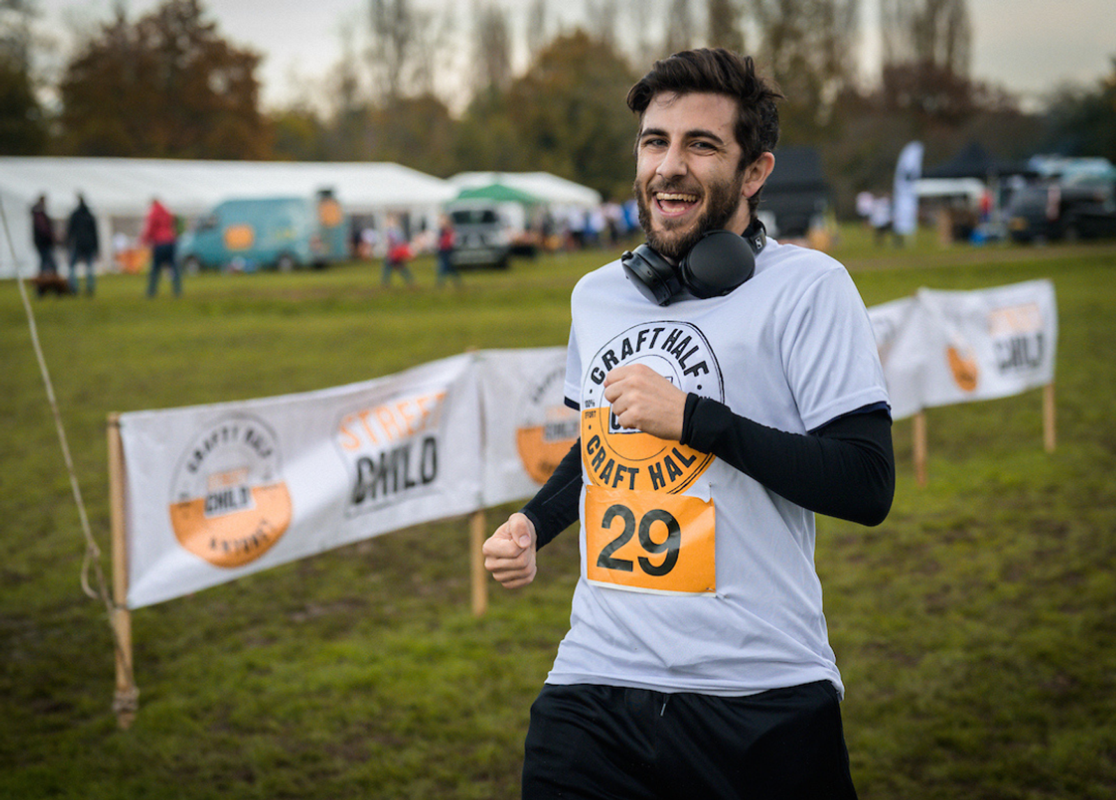 A man is running in an outdoor race, wearing a white "GRAFT HALF" shirt with a number 29 bib and headphones around his neck. In the background, there are banners that read "Street Child" and "LOCAL," along with multiple participants and tents on grassy terrain.