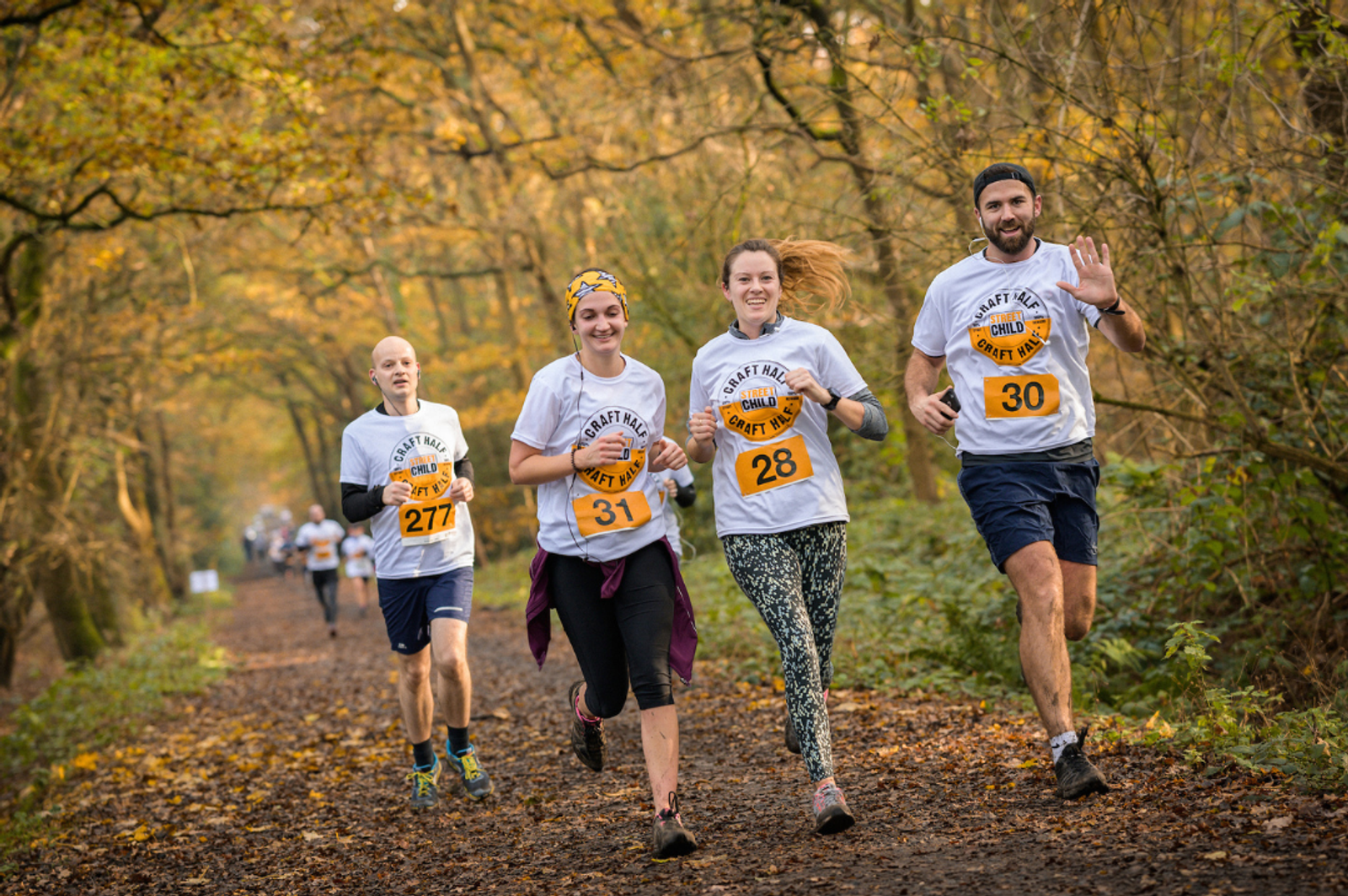 Four runners wearing numbered bibs and matching white shirts run along a leaf-covered forest trail during the Craft Half event for Street Child. They appear in high spirits, with one runner raising both thumbs up. A "LOCAL" logo is displayed at the bottom of the image.