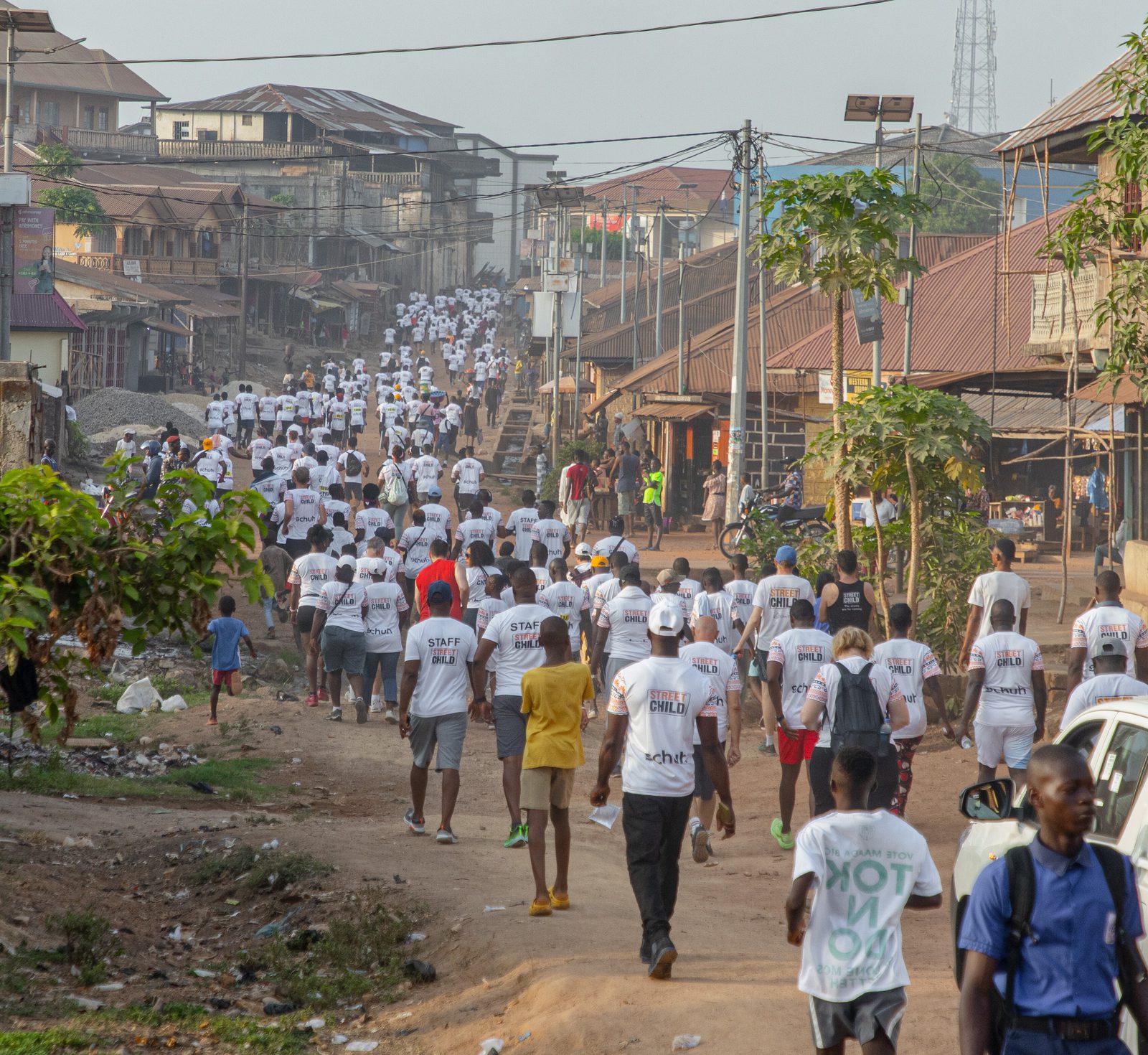 A large group of people wearing white t-shirts walk down a dirt road in a residential area. Some buildings, utility poles, and scattered debris are visible. The t-shirts display various logos and text. Onlookers stand by as the crowd moves forward.