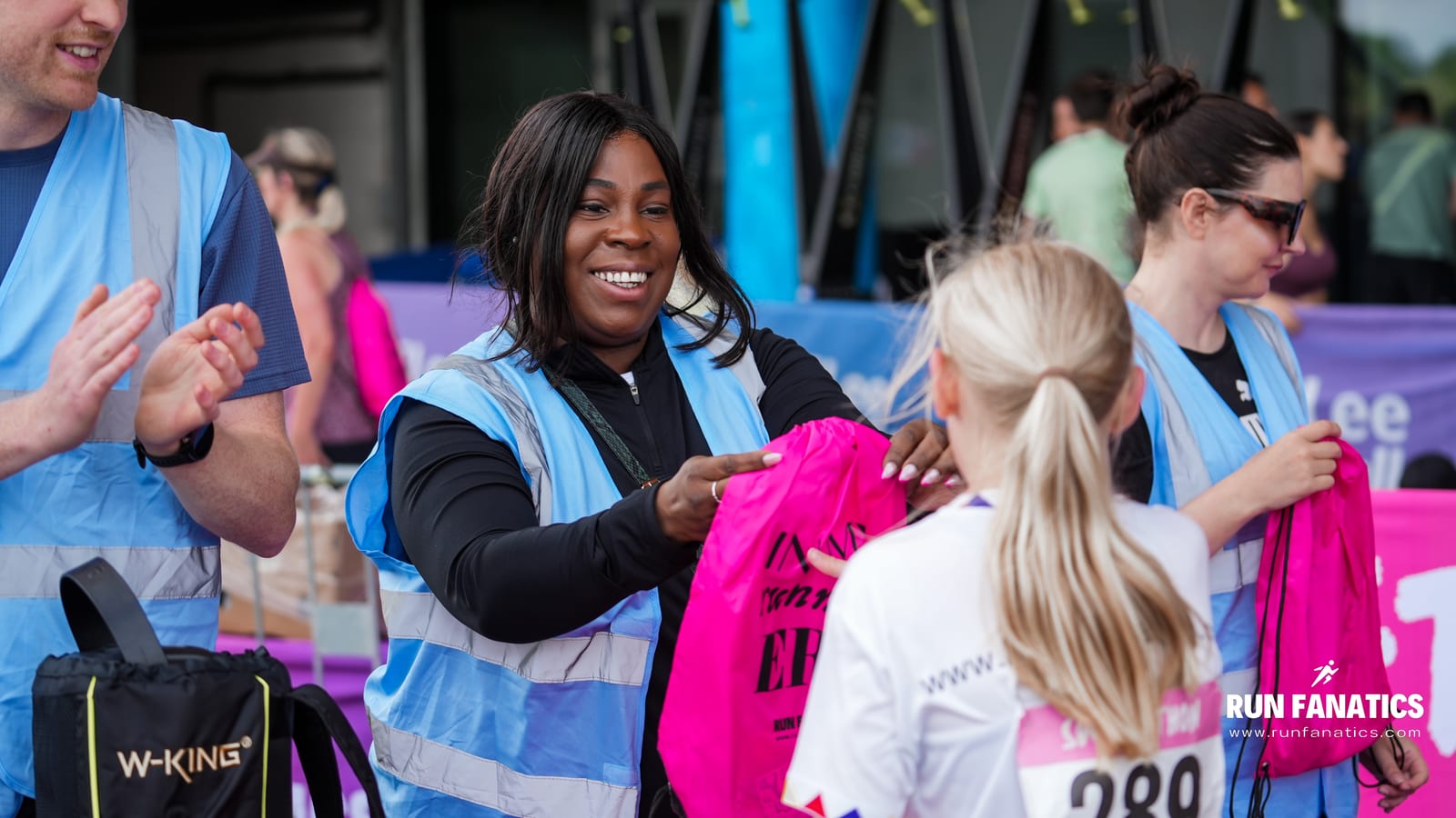 A smiling volunteer in a blue vest hands a pink bag to a participant at a running event, while others assist nearby. "RUN FANATICS" branding is visible in the corner.
