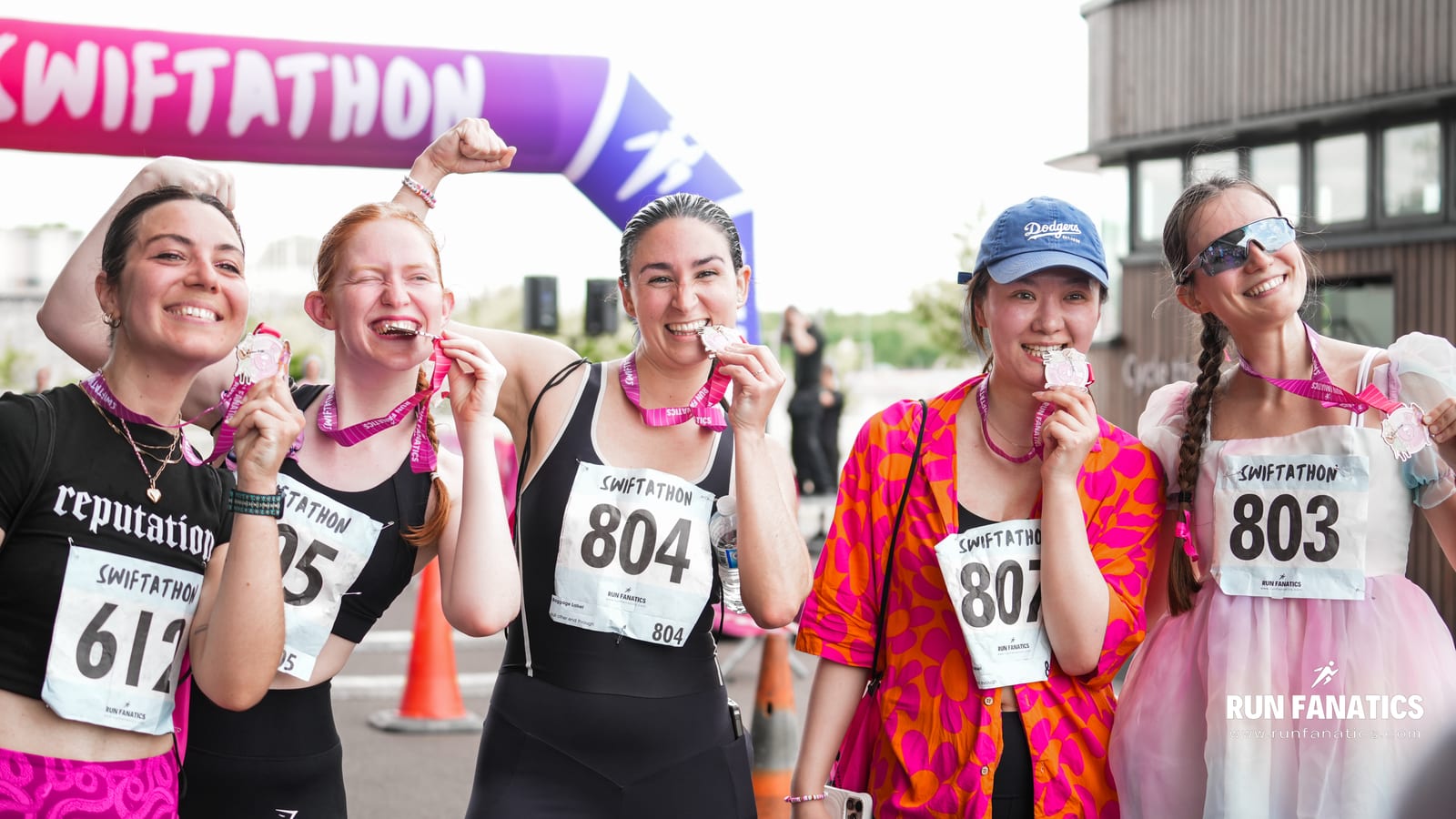 Five smiling women pose after a race, each wearing medals and race bibs. They stand in front of a colorful "Swiftathon" banner, celebrating their achievement, some playfully biting their medals.