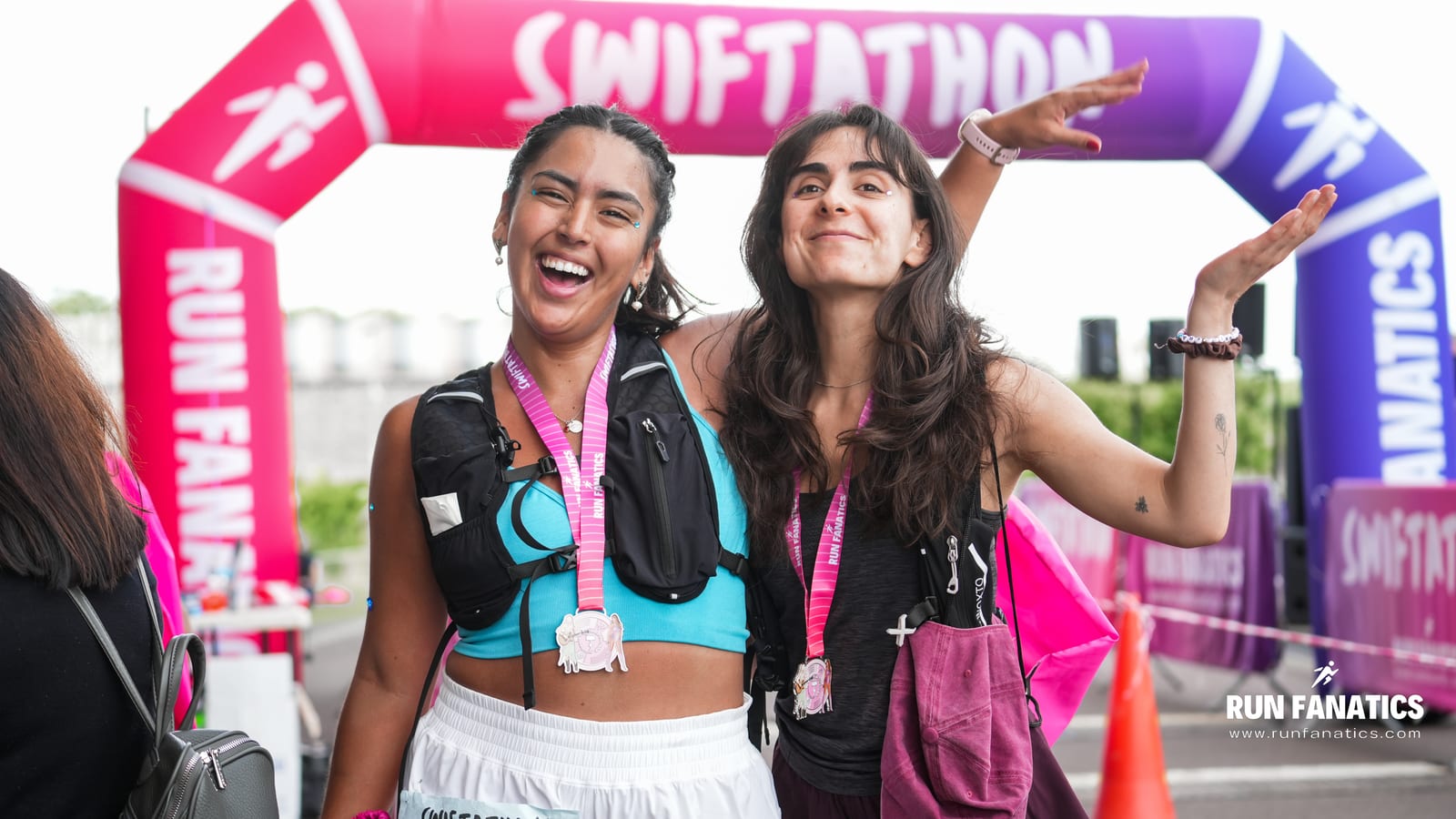 Two smiling women wearing medals and race bibs pose happily in front of a colorful "Swiftathon" finish line arch at a running event. Both wear athletic gear; one gestures playfully with her arm. The atmosphere is celebratory.