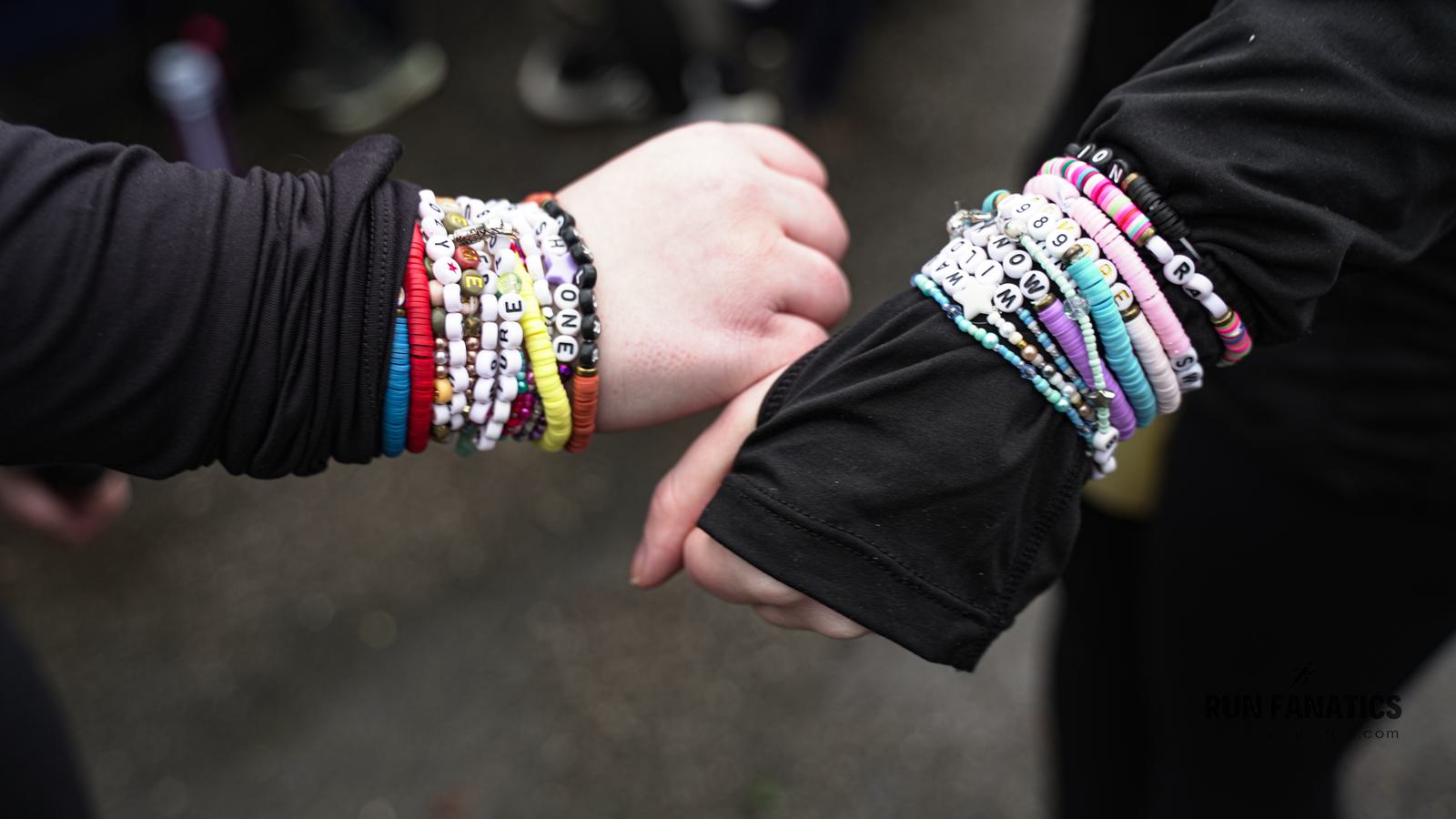 Two people wearing black sleeves touch fists. Both wrists are adorned with numerous colorful beaded bracelets featuring letters and symbols. The background is blurred, highlighting the focus on the hands and bracelets.