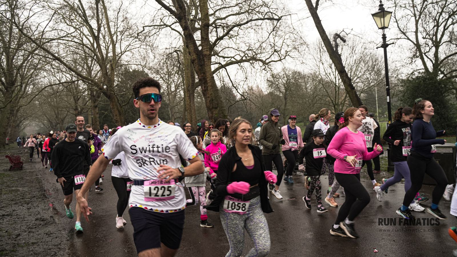 A group of people running in a park during a race event. Participants wear numbered bibs, and some have themed shirts. Trees without leaves line the path, and spectators are visible in the background. It's a cloudy day.
