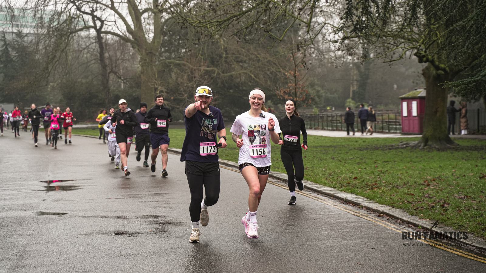 Runners participate in a race on a wet, overcast day in a park. A man and woman in front smile and gesture, wearing numbered bibs. Other runners follow behind on the asphalt path lined with grass and trees.