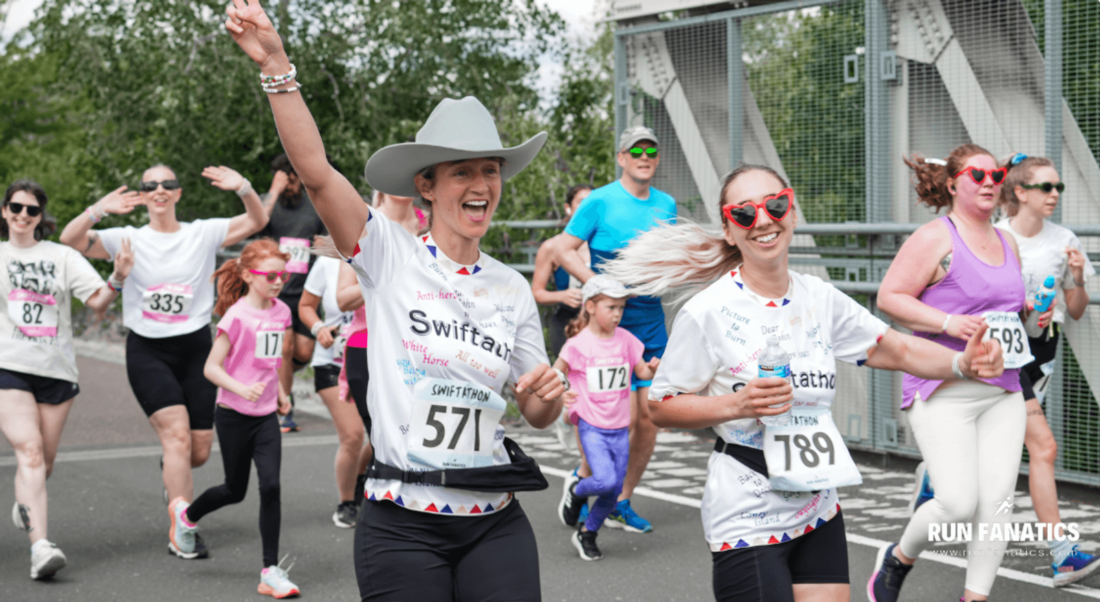 A group of smiling runners, many wearing sunglasses and race bibs, participate in a fun run. Two women in front, one in a large gray cowboy hat, wave and smile. Other runners and children follow behind on a bridge.
