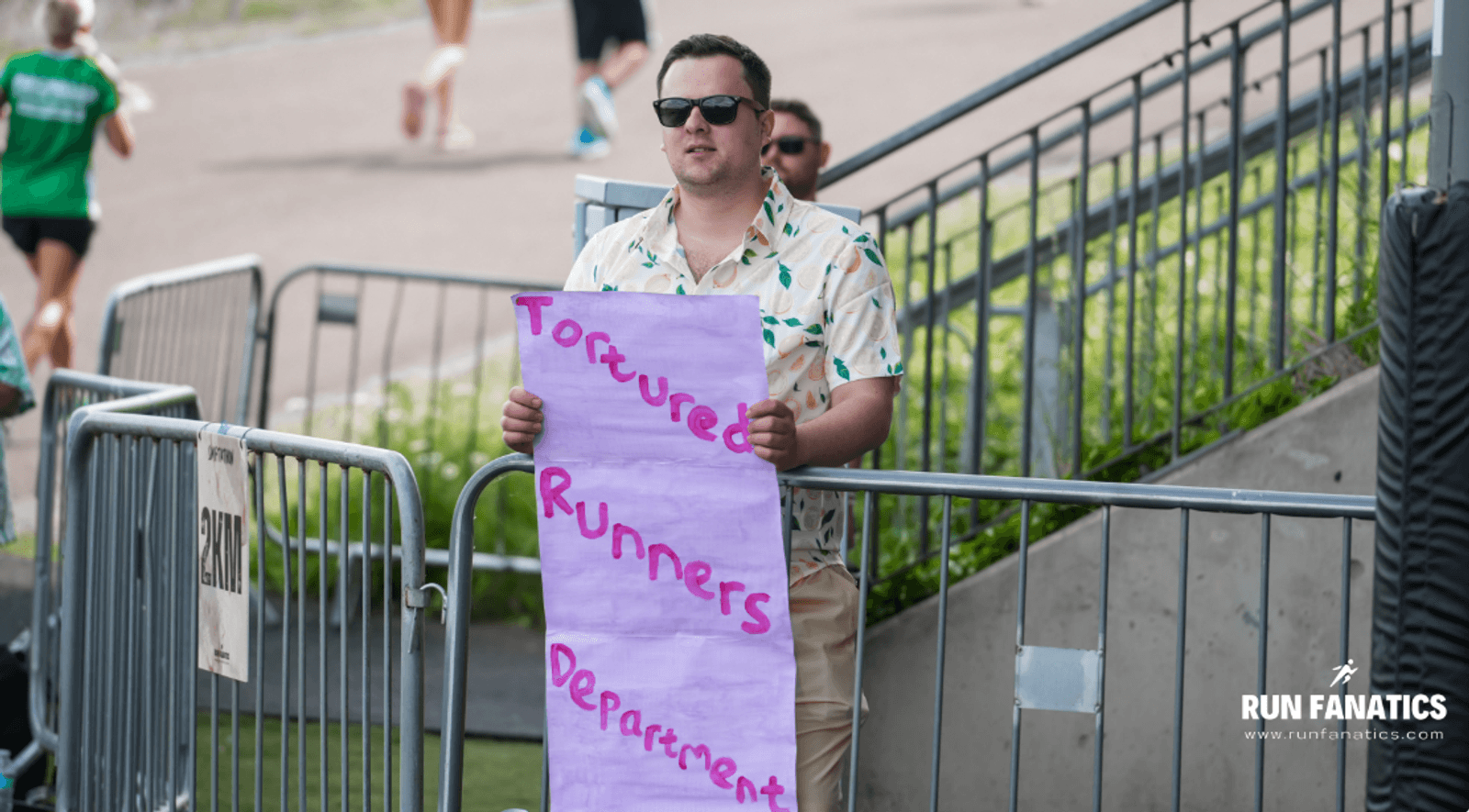 A man in sunglasses and a patterned shirt holds a handwritten purple sign that says "Tortured Runners Department" near a race barrier at an outdoor running event. The Run Fanatics logo is visible in the corner.