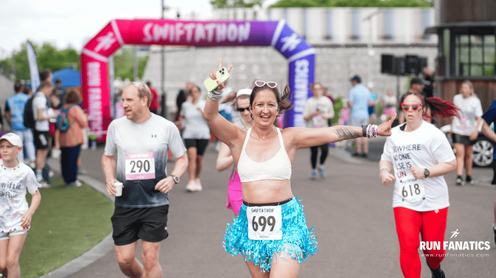 A smiling woman wearing a race bib 699 and a blue tinsel skirt waves while running in a race, surrounded by other runners near a colorful inflatable arch that reads "SWIFTATION.