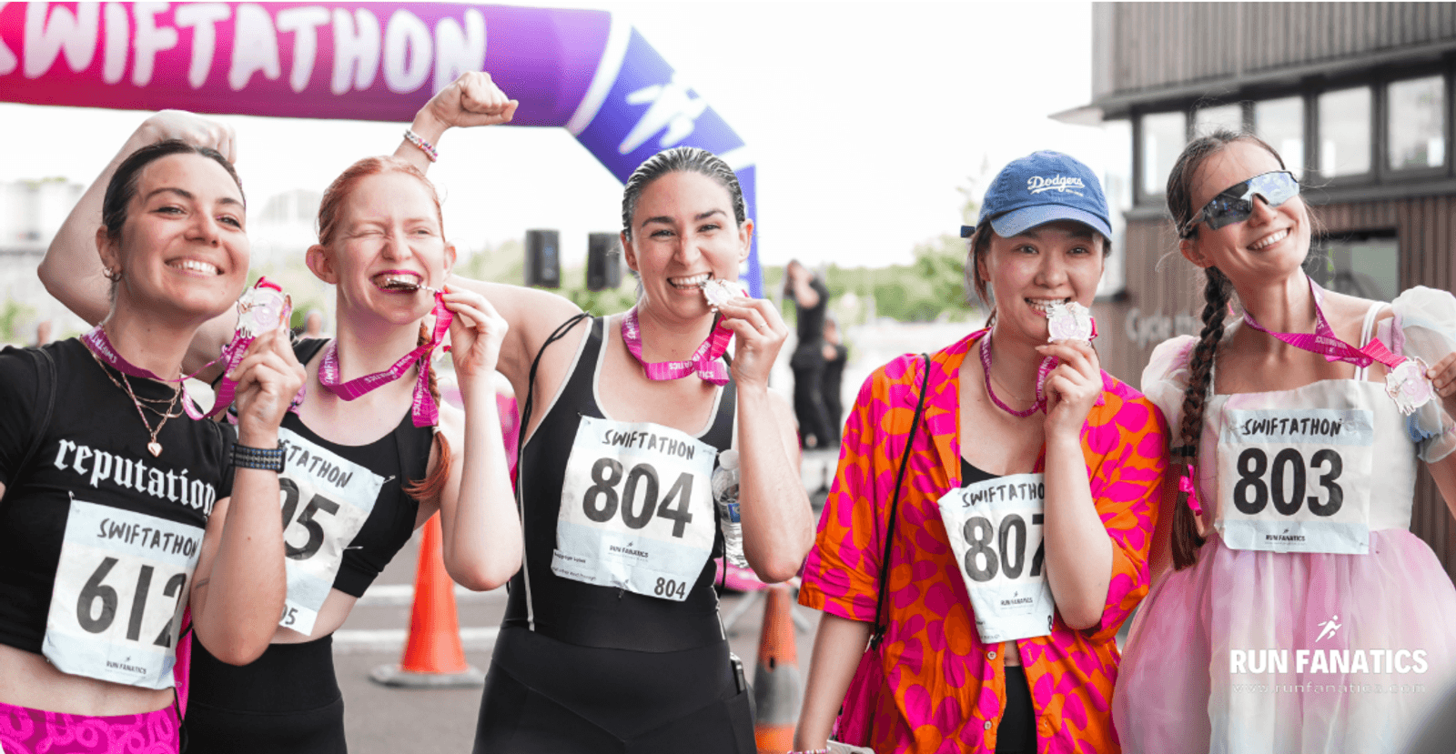Five smiling runners with race bibs pose at a finish line, playfully biting their medals. They wear colorful outfits and “SWIFTATHON” bibs, celebrating after completing an event. The mood is joyful and victorious.