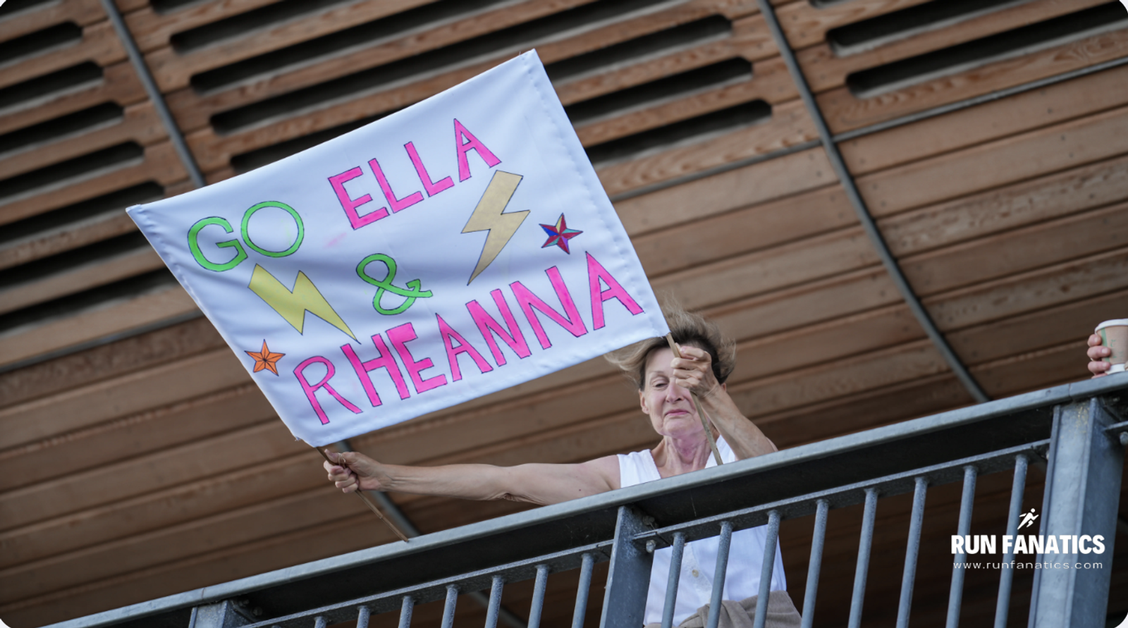 A person on a balcony holds a handmade sign that reads "GO ELLA & RHEANNA" with colorful letters and lightning bolt drawings. The background shows wooden paneling and a "Run Fanatics" logo.