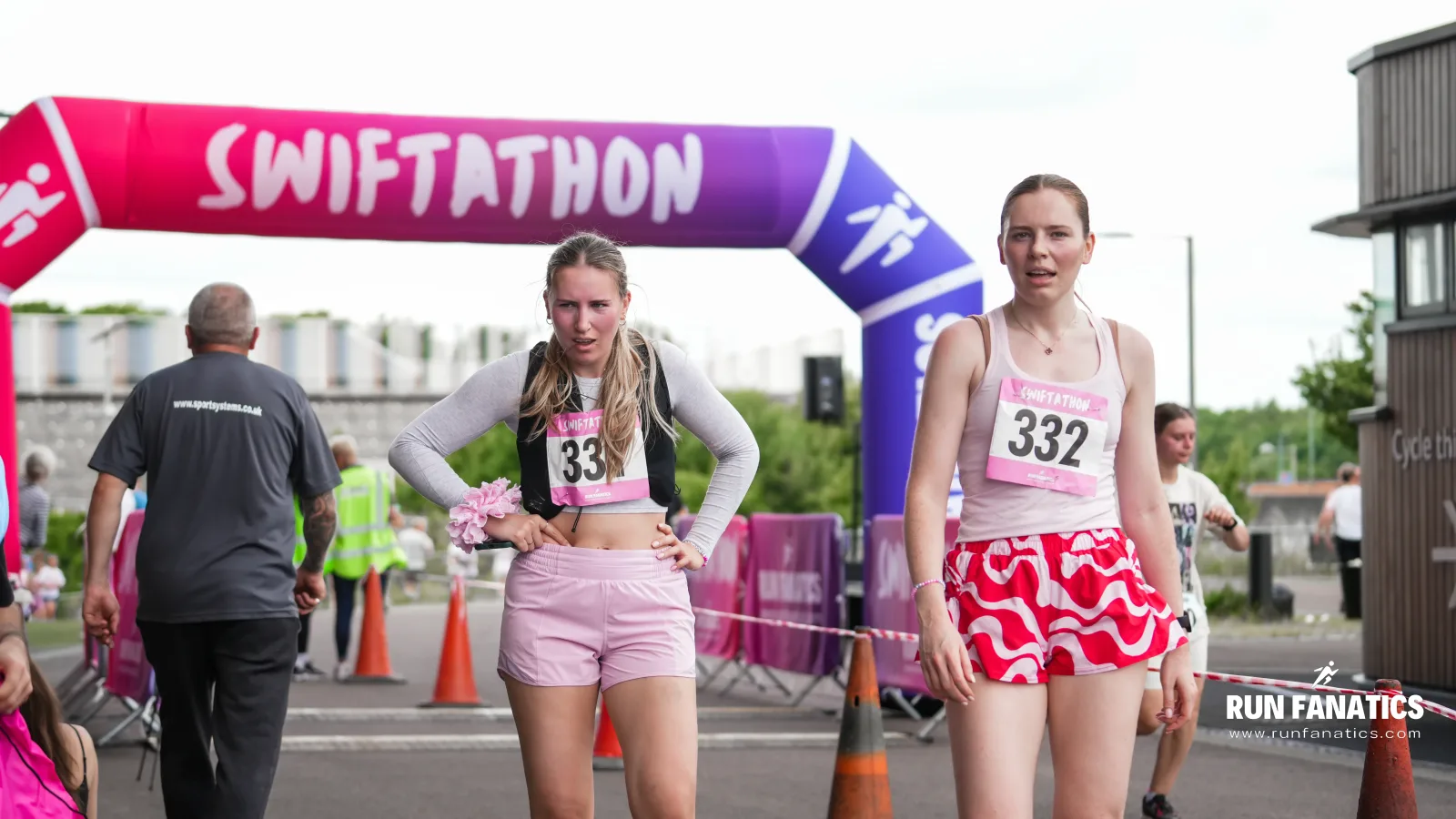 Two women wearing race numbers 331 and 332 stand near a finish line arch labeled "SWIFTATHON." Both appear tired after running. Other people and race signage are visible in the background.