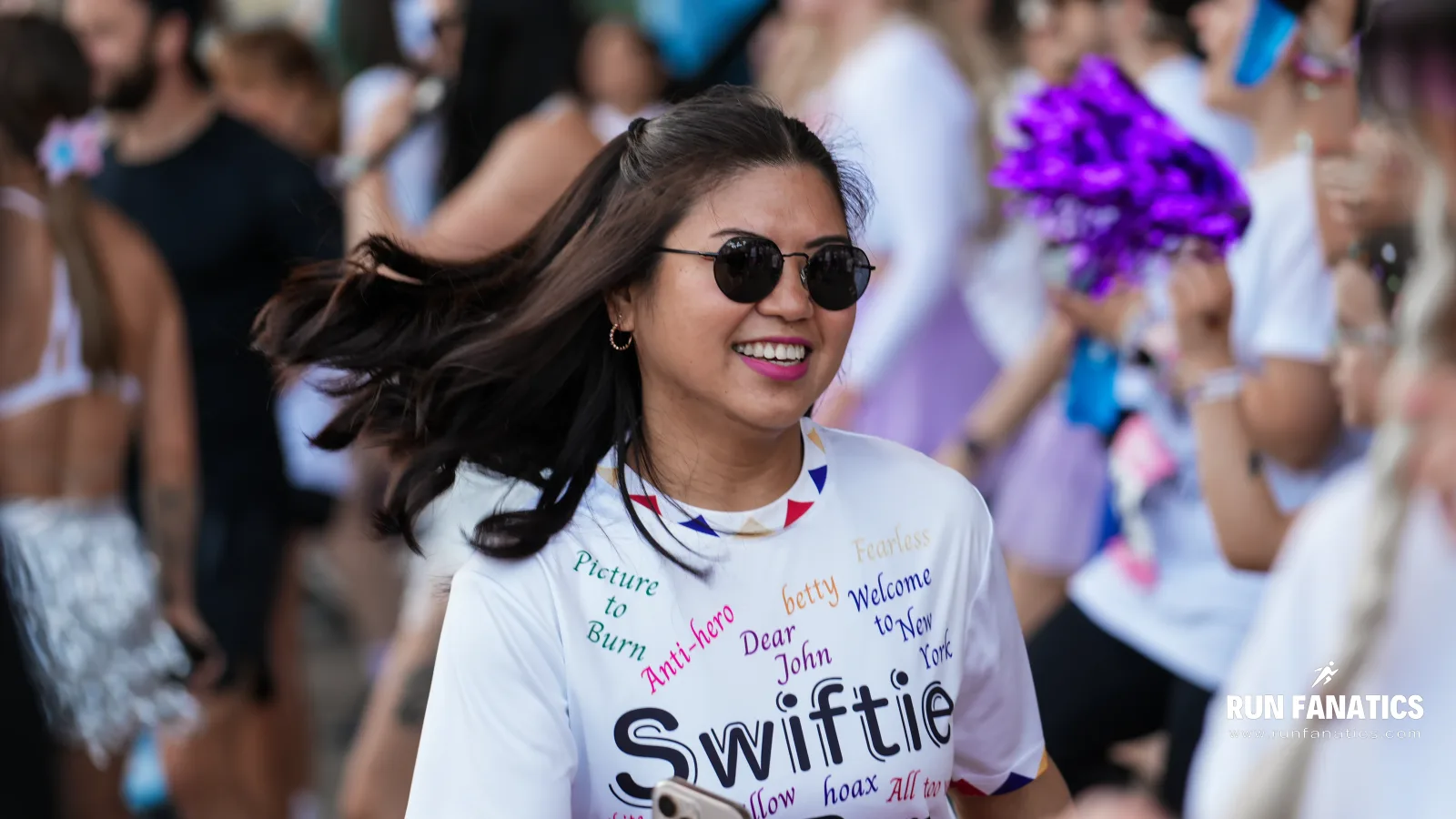 A woman wearing sunglasses and a "Swiftie" T-shirt smiles while running in a crowded outdoor event. People in festive clothing and a cheerleader with purple pom-poms are visible in the background.
