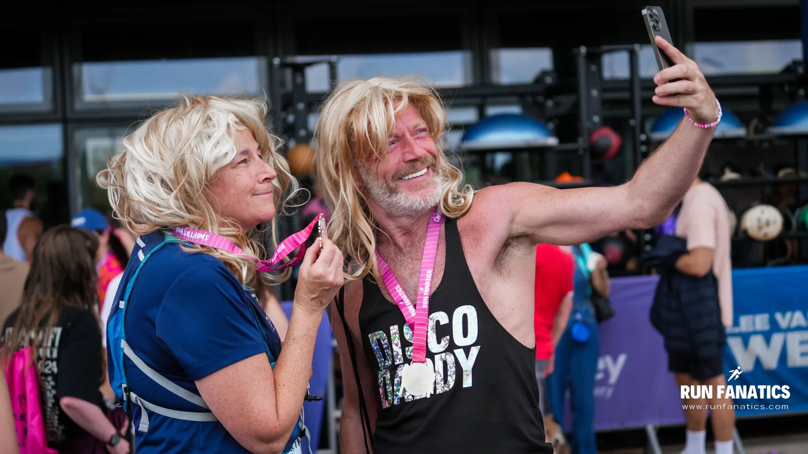 Two people wearing blonde wigs and finisher medals smile and pose for a selfie at a running event. One wears a blue shirt and the other a black "DISCO DOLLY" tank top. "Run Fanatics" logo is visible in the corner.