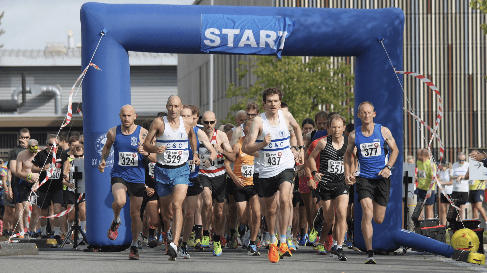 A group of runners is starting a race, all dressed in athletic gear, under a blue inflatable arch labeled "START." Some are wearing bib numbers. The background shows industrial buildings and a clear sky.