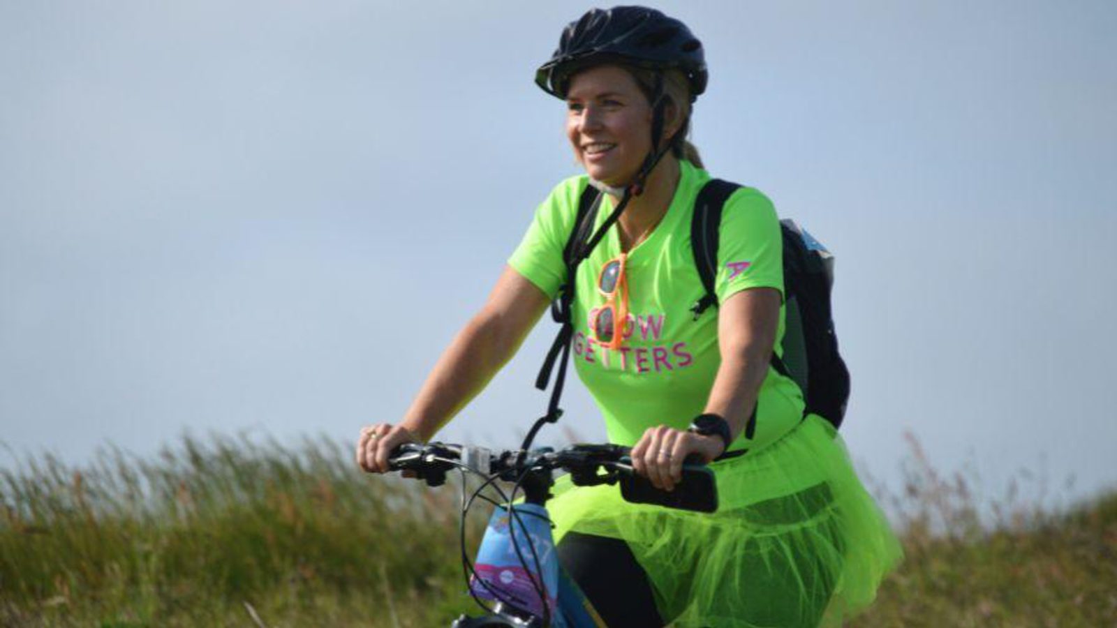 A woman wearing a bright green shirt and matching tutu rides a bicycle outdoors. She is smiling, wearing a helmet and sunglasses, and there is grass and sky in the background.