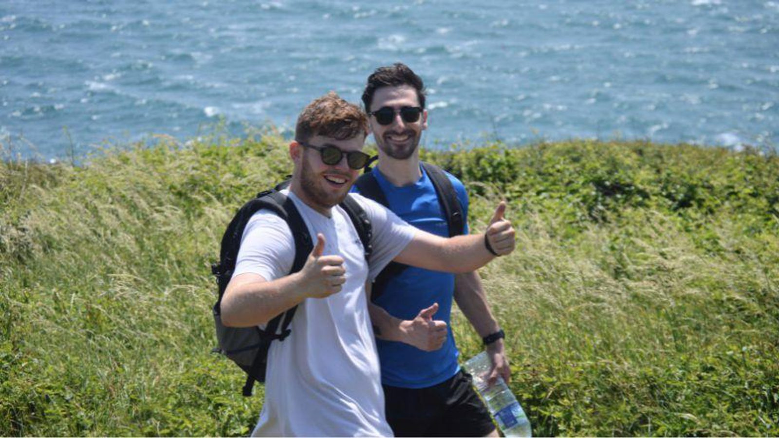 Two men with backpacks smile and give thumbs up while standing on grassy terrain near the ocean on a sunny day. One holds a bottle of water.