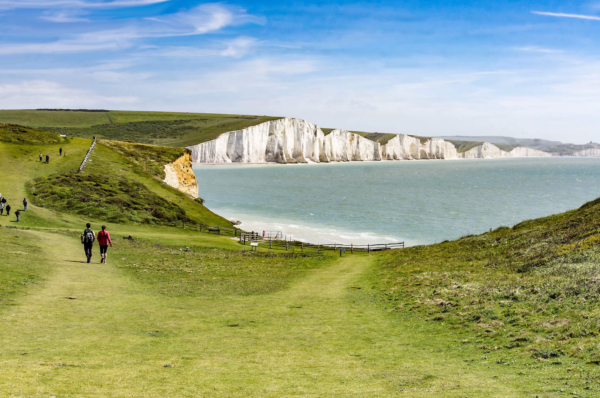 People walk along a grassy path on a coastal cliffside, with white chalk cliffs and the blue sea visible in the distance under a bright, partly cloudy sky.