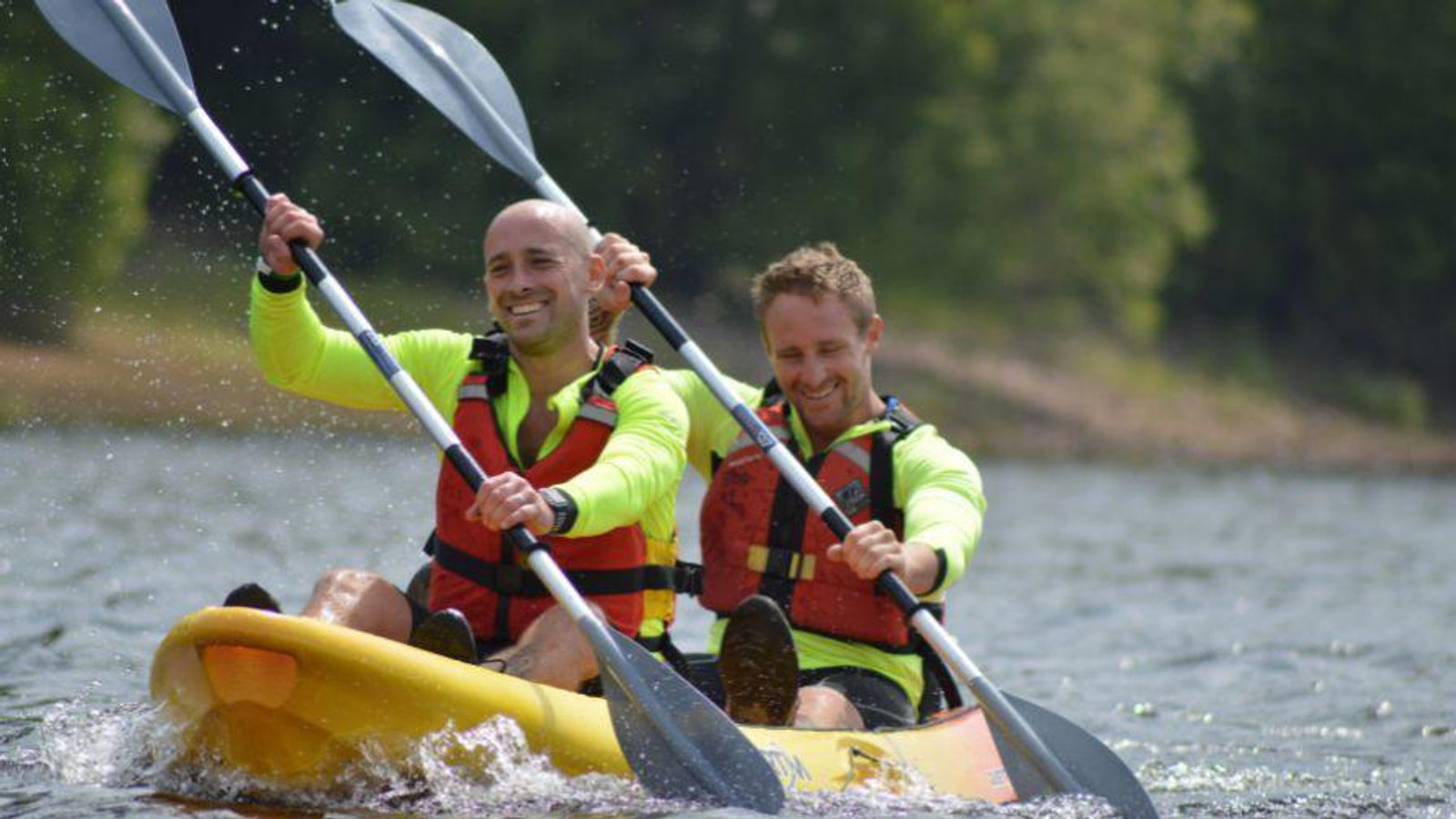 Two men wearing life jackets and neon green shirts paddle a yellow kayak together on a lake, smiling as they move through the water with trees visible in the background.