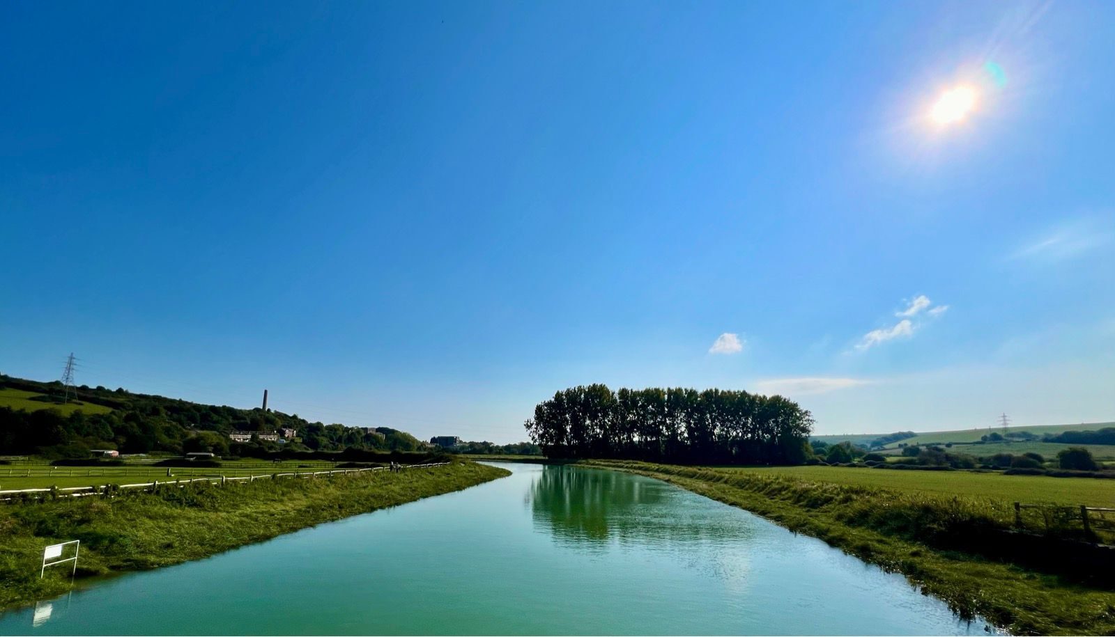 A serene river flows through a verdant countryside under a clear blue sky with the sun shining brightly. Trees line the river's edge in the distance, and lush green fields stretch out on either side.