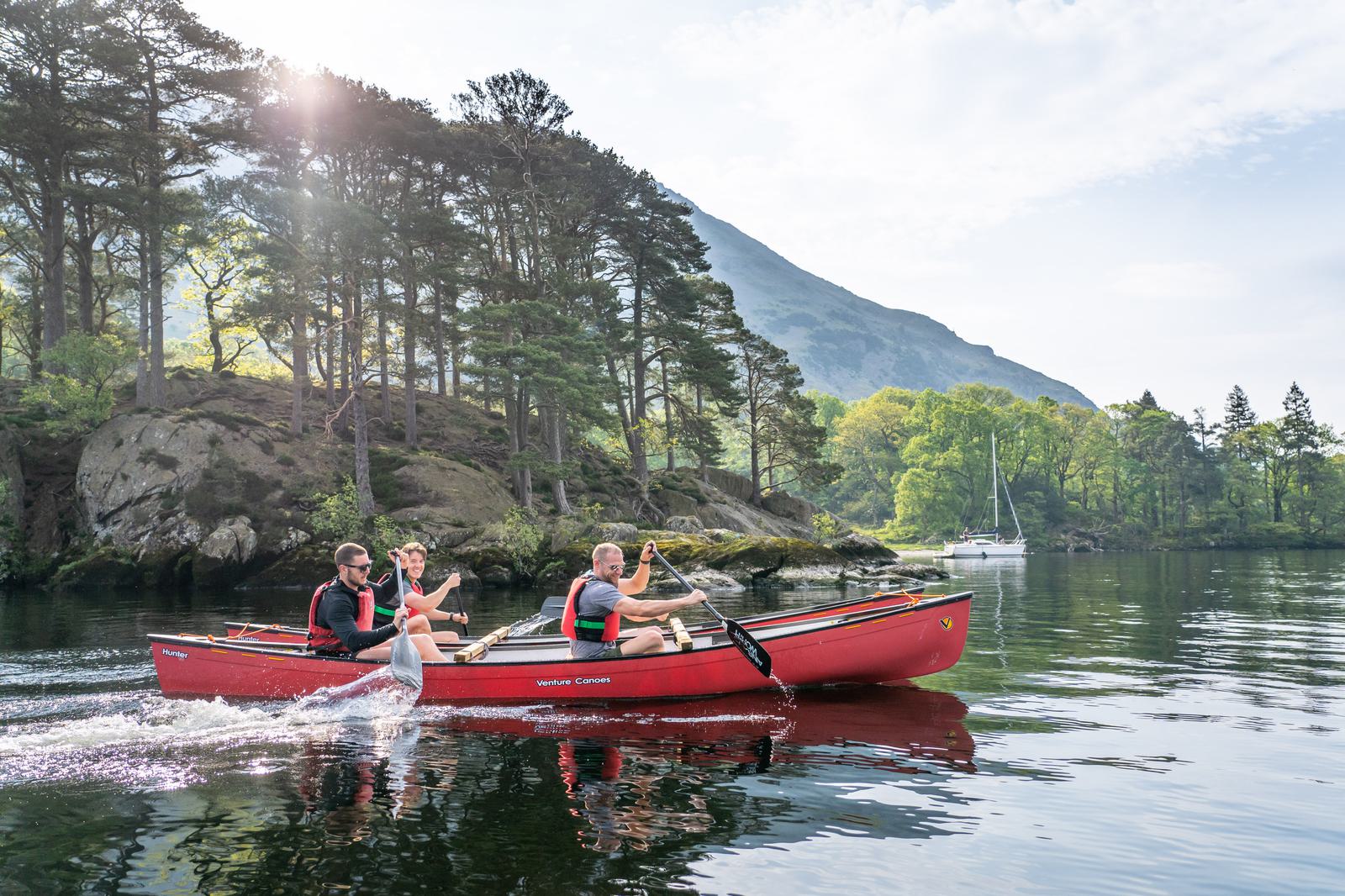 Three people in a red canoe paddle on a calm lake surrounded by lush greenery and a mountain in the background. The sun shines through the trees, creating a serene and picturesque outdoor setting.