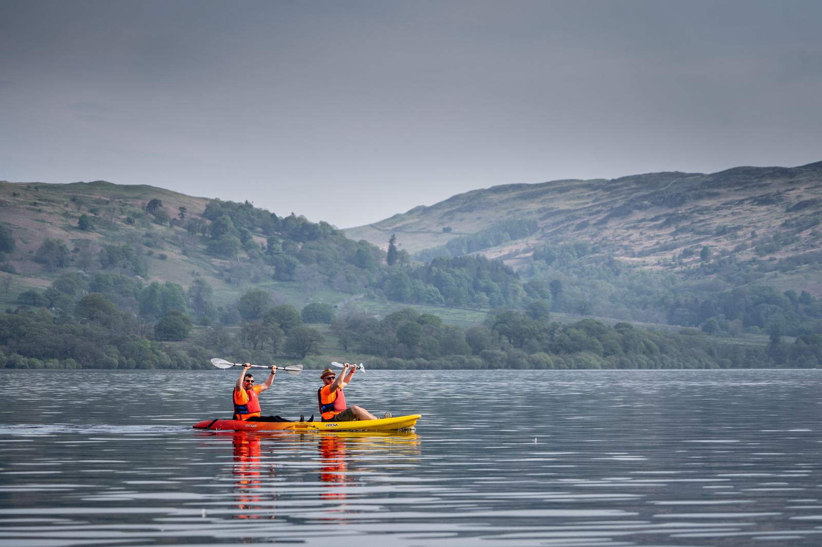 Two people in brightly colored kayaks paddle on a calm lake surrounded by green, rolling hills under a cloudy sky. Both are wearing orange life jackets and appear to be enjoying a serene outdoor adventure together.