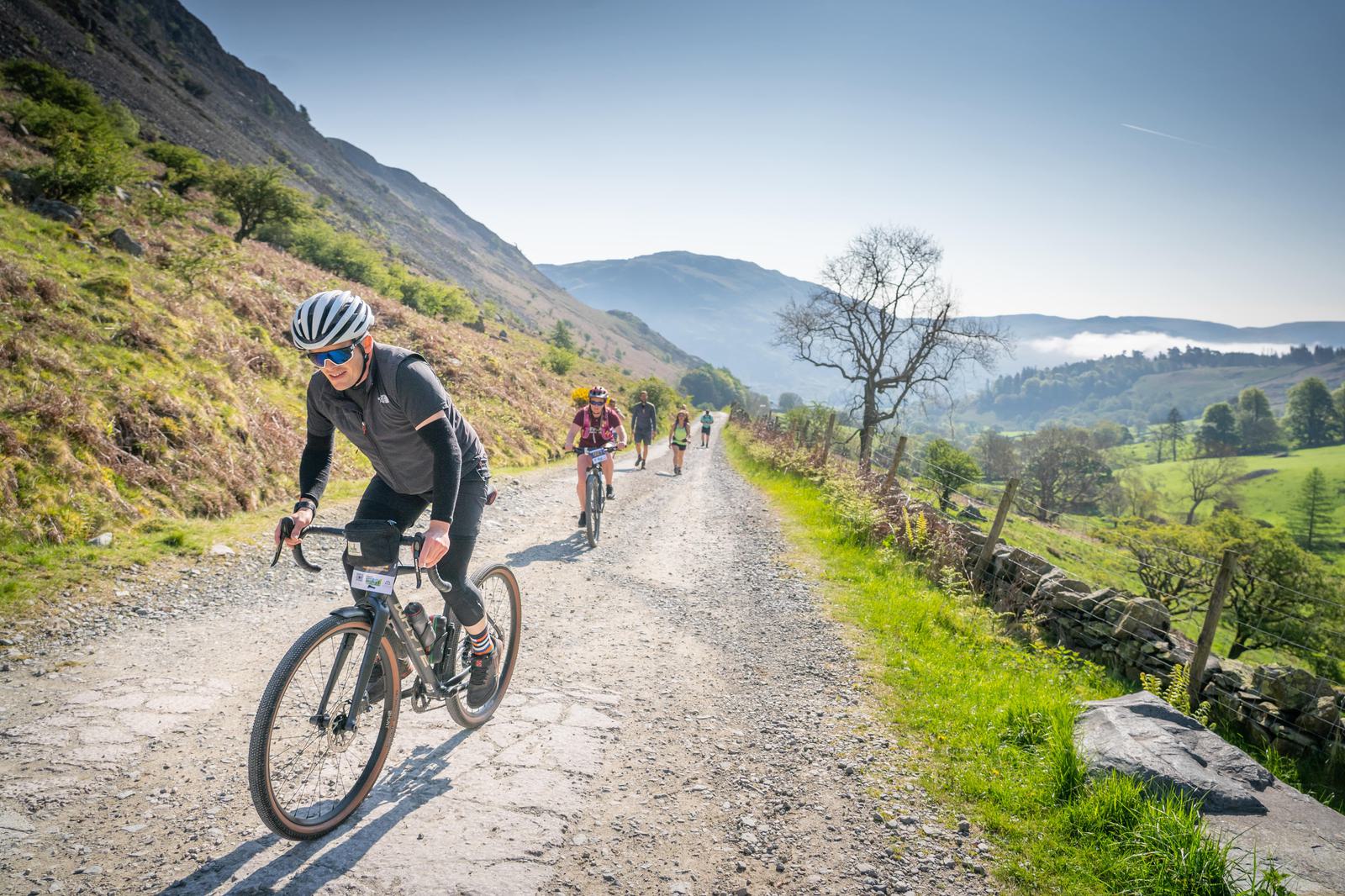 A group of cyclists rides along a scenic, gravel mountain path on a sunny day, surrounded by lush green fields and rolling hills. The lead cyclist, wearing a helmet and sunglasses, concentrates on the trail ahead.