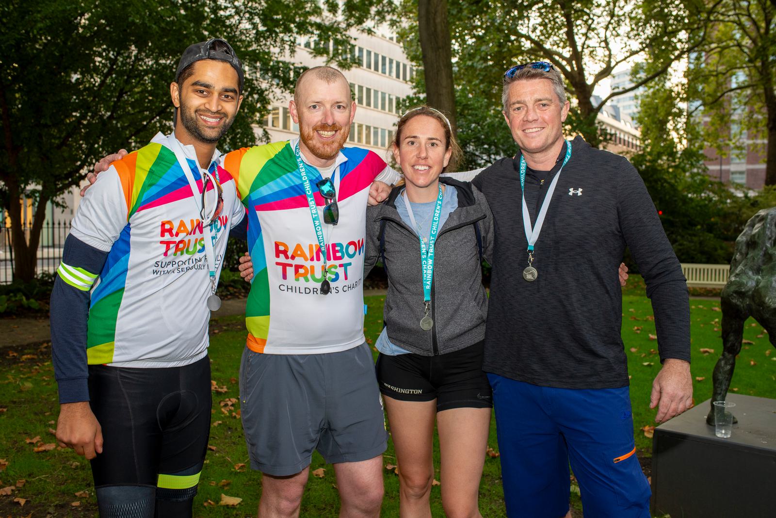 Four cyclists with medals pose for a photo in a park. Two are wearing "Rainbow Trust Children's Charity" jerseys. They stand with arms around each other, smiling. Trees and a building are visible in the background.