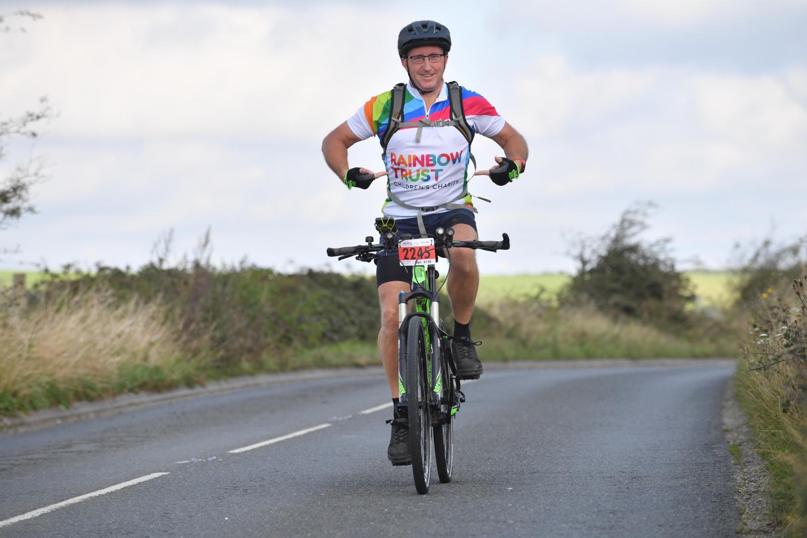 A cyclist wearing a colorful jersey and helmet rides a bicycle on a rural road. The jersey has "Rainbow Trust" written on it. The cyclist is smiling, and the scenery includes grass and cloudy skies. A race number is visible on the bike.